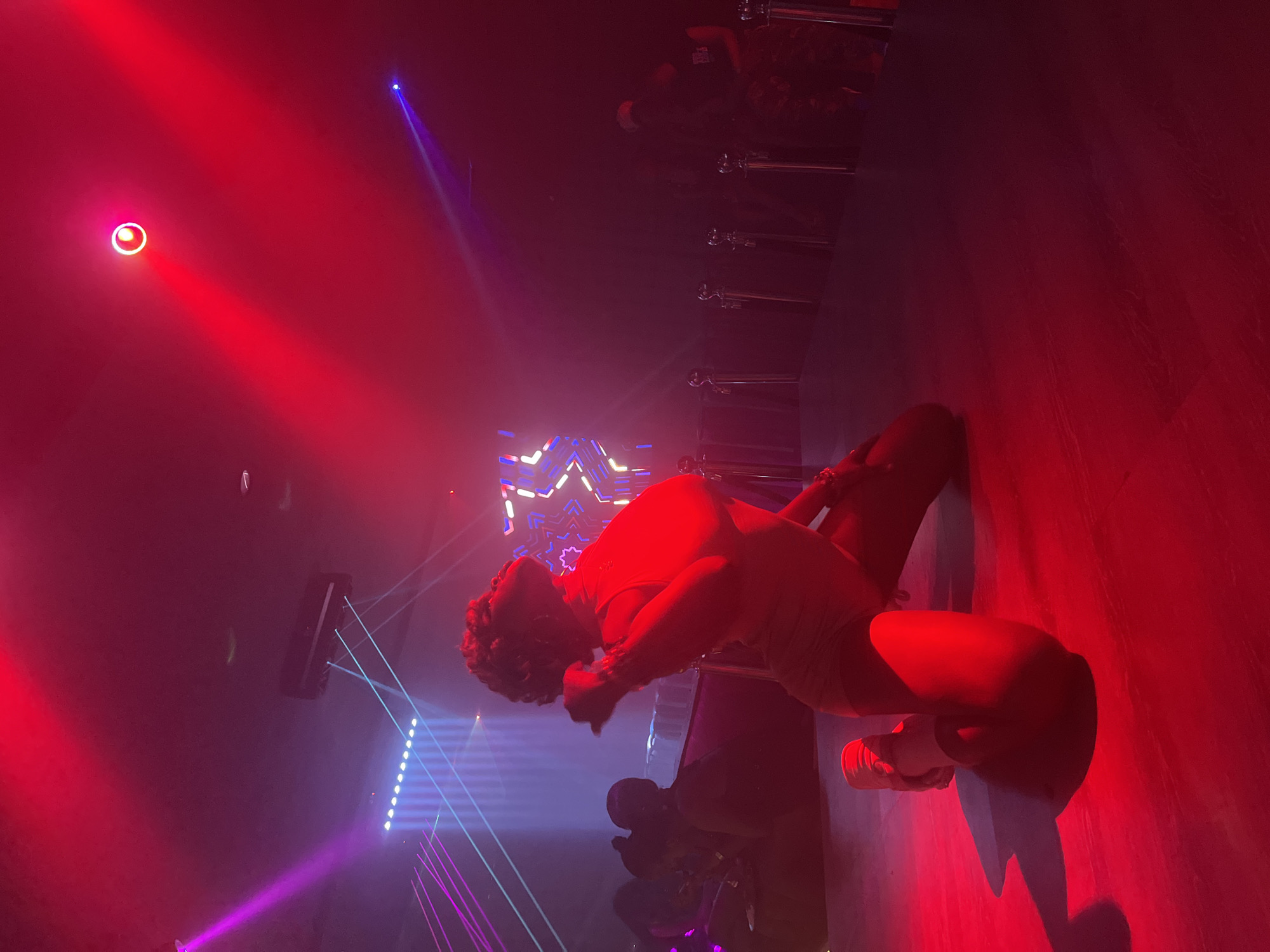 The image depicts a dimly lit interior, likely a club or performance venue in Howayi, Nigeria. The dominant lighting is red, casting a warm glow over the scene. In the foreground, a person is positioned on their knees, facing away from the camera and looking upwards. They are wearing a dark-colored garment that appears to be a dress or leotard. The person's pose suggests they are either performing or engaging in an activity that draws attention to their upper body and head.
The background is blurred but reveals elements of a stage or performance area. Several rows of stanchions with ropes are visible, indicating a roped-off area, possibly for VIPs or a stage perimeter. Further back, faint silhouettes of other individuals can be seen, some seated and others standing. Above, lighting fixtures are present, including what appears to be a moving head light with a red beam and a circular spotlight. There are also illuminated panels with geometric patterns in the background, contributing to the atmospheric lighting. The floor is a dark, reflective surface, with shadows cast by the foreground subject and the lighting. The overall ambiance is one of nightlife and entertainment.