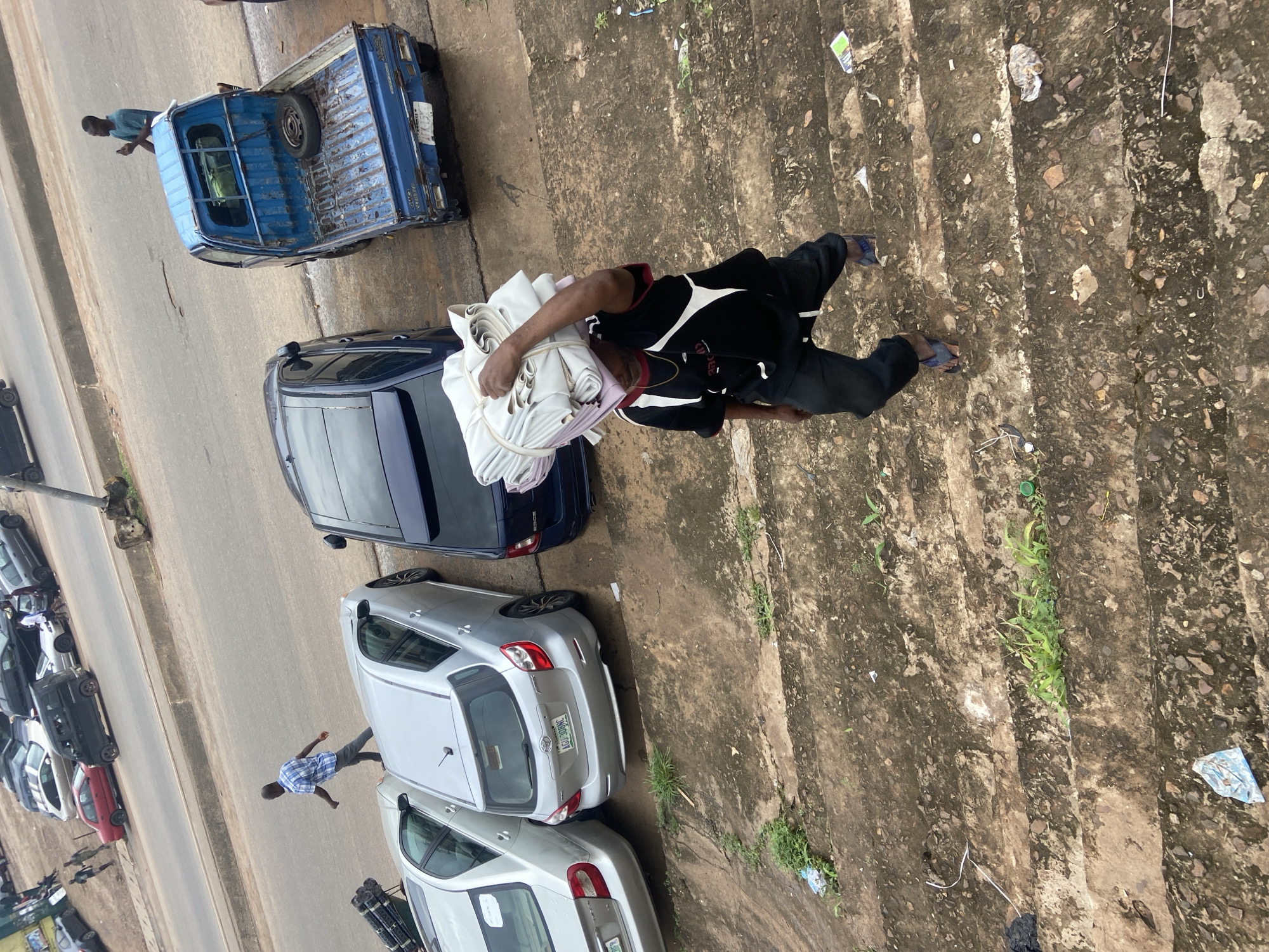 The image, taken from an elevated perspective in Ogbunike, Nigeria, shows a street scene with several cars parked along the side of a road. A blue pickup truck is parked on the left side of the frame, with a spare tire visible in its bed. A person in a blue t-shirt is standing in the truck bed, looking down. A man in a black and white shirt and black pants is walking on a rough, textured surface, carrying a bundle of white fabric. Several cars are parked on the road, including a dark blue sedan, a silver hatchback, and other vehicles further down the street. The ground is made of concrete with some patches of dirt and grass growing through cracks. There is a noticeable amount of debris and litter scattered on the ground. The lighting suggests it is daytime, but the weather appears overcast. The overall impression is of a busy, working-class area.