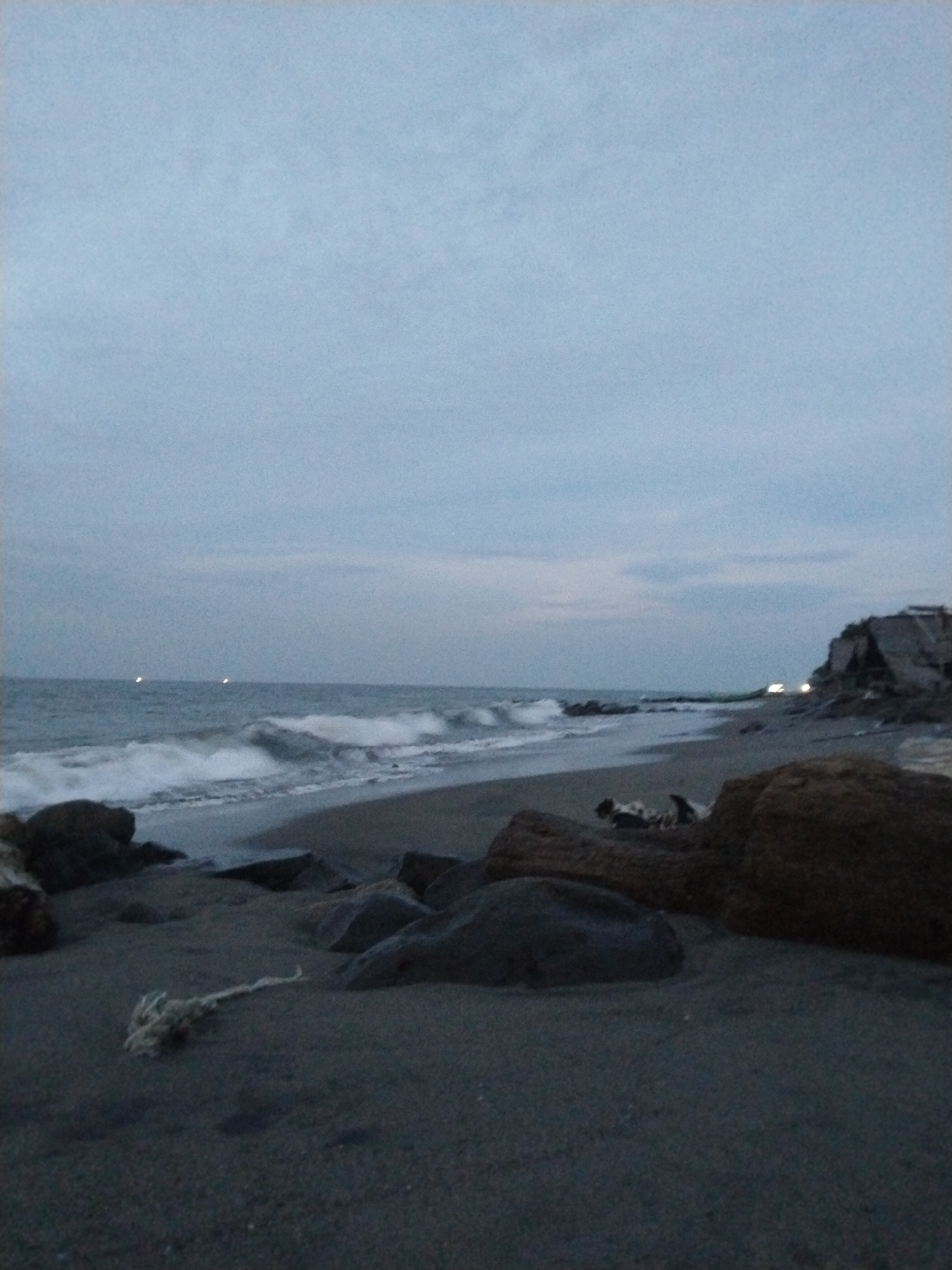 The image depicts a beach scene at dusk or dawn. The sky is overcast with muted blue and gray tones, with some indistinct clouds. The sea is visible in the background, with moderate waves breaking on the shore. The beach itself is composed of dark sand and scattered rocks, some of which are quite large. In the foreground, there is a piece of white rope or netting lying on the sand. In the distance, there appear to be a few small lights on the horizon over the water, possibly from boats or structures. On the far right side of the image, there is a rocky outcrop or structure with some debris. The scene suggests a natural environment, likely a coastal area. Without further information or context, it is not possible to determine the exact location within Lhokseumawe, Indonesia, or to identify specific people, actions, or interactions.