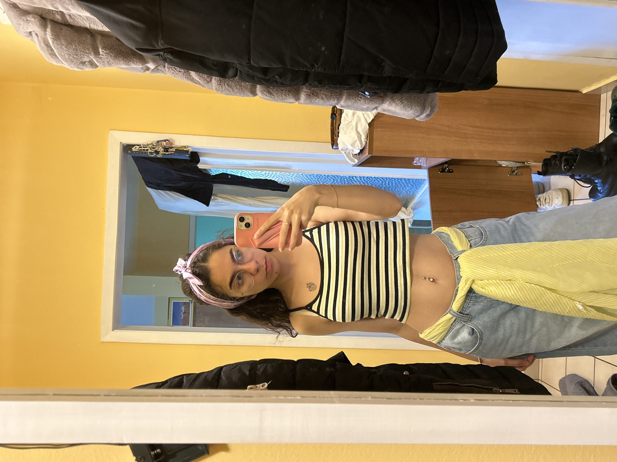 A woman is taking a selfie in a mirror. She is wearing a striped crop top, jeans with a yellow scarf tied around her waist, and a pink headband. She has glasses on and is holding a pink phone. The room has yellow walls and a wooden dresser. There are clothes hanging on the wall. A picture can be seen on the wall in the background. The location appears to be indoors. The location in the city of Sofia, Bulgaria cannot be determined from the image.