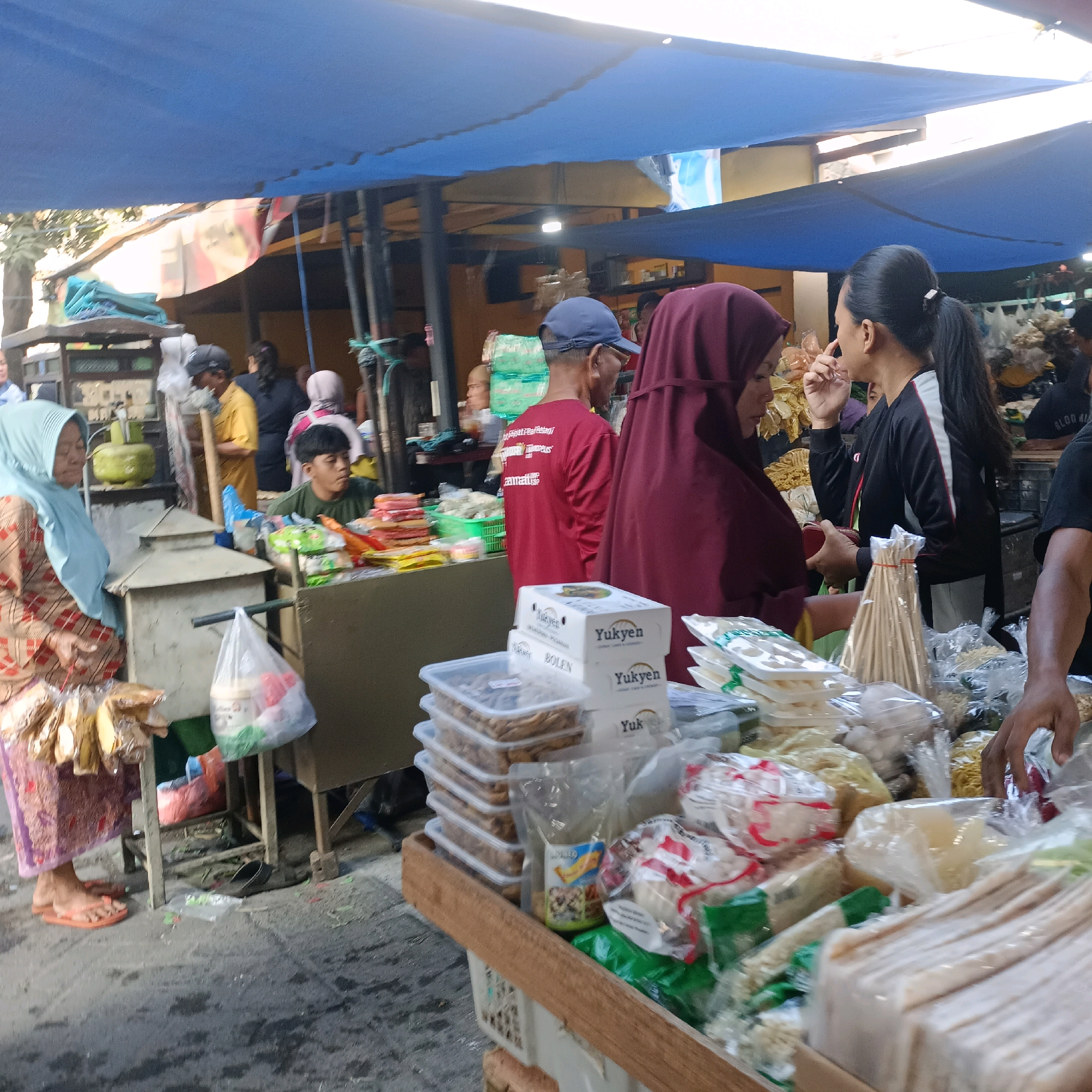The image depicts a street food stall in Surabaya, Indonesia, likely a market or busy street. The scene is bustling with activity, with several people present. In the foreground, a woman wearing a light blue hijab and a patterned dress is at the stall. Next to her, a man in a red shirt and a blue cap is also present. Further back, other individuals are visible, including a woman in a maroon hijab and a woman with her hair in a ponytail.
The stall is laden with various food items, many of which are packaged in plastic. There are stacks of what appear to be snacks or dried goods in clear plastic containers, bags of noodles, and other packaged food products. A green gas cylinder is visible behind the stall. The entire setup is covered by a large blue tarp, providing shade from the sun. The ground is paved with asphalt, showing some wear and tear. The overall impression is of a vibrant and active marketplace in a city.