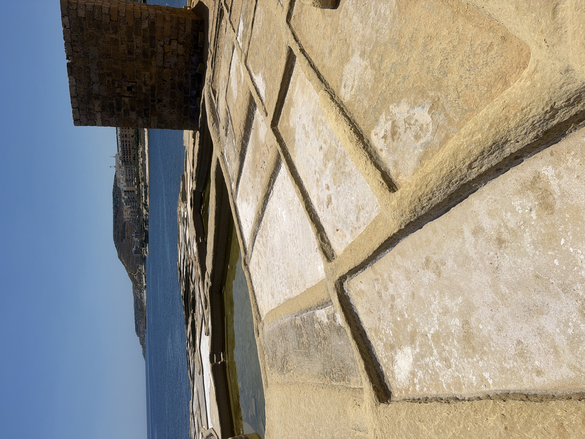 The image depicts a salt pan in Marsalforn, Malta, under a clear blue sky. In the foreground, the salt pans are carved into the rocky terrain, forming a grid of shallow rectangular pools. Some pools contain a shallow layer of greenish-brown water, while others are dry and appear to have a white, crystalline residue, indicative of salt. The surrounding rock is a warm, sandy yellow. To the right, a weathered, square stone tower stands prominently, constructed from irregular, light brown stones. In the background, the deep blue Mediterranean Sea stretches to the horizon. Beyond the sea, a coastal town with buildings and hills is visible, suggesting a populated area. The scene is devoid of people.