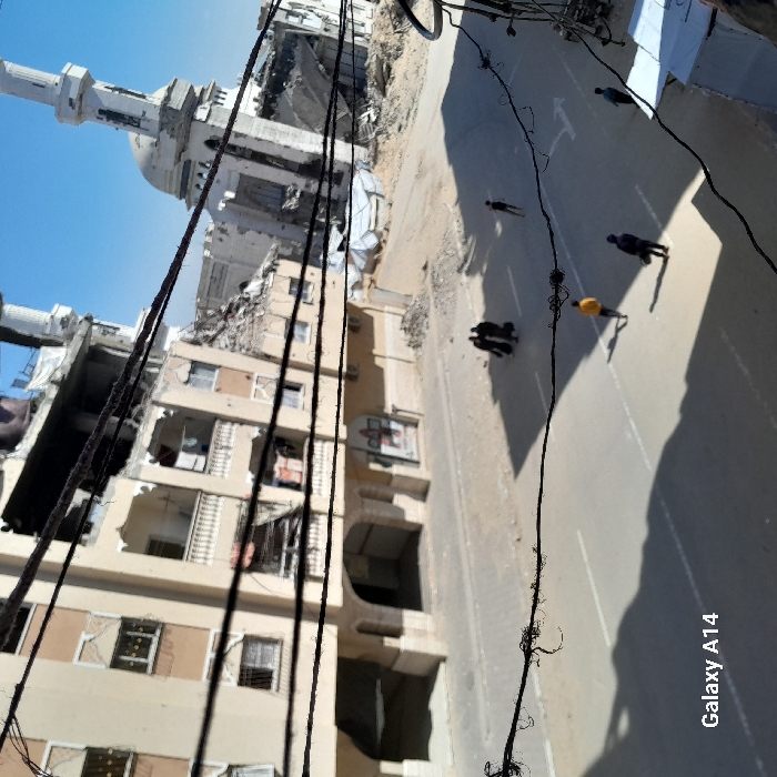 The image depicts an urban street scene in Khan Yunis, Palestinian Territory, under a clear blue sky. Numerous tangled black wires crisscross the foreground, partially obscuring the view. To the left, a multi-story, light-colored building exhibits significant structural damage, including collapsed sections, exposed interiors, and rubble piled at its base. To the right, in the background, a mosque with a dome and two minarets is extensively damaged, with large portions of its structure missing and surrounded by considerable debris. The paved street traversing the midground features white road markings. Six individuals are visible on the street: a group of three in dark clothing walks on the left, while three other individuals, including one in a yellow top, are spread further right, either walking or standing. Long shadows are cast across the street, indicating daytime. The environment conveys widespread structural devastation.