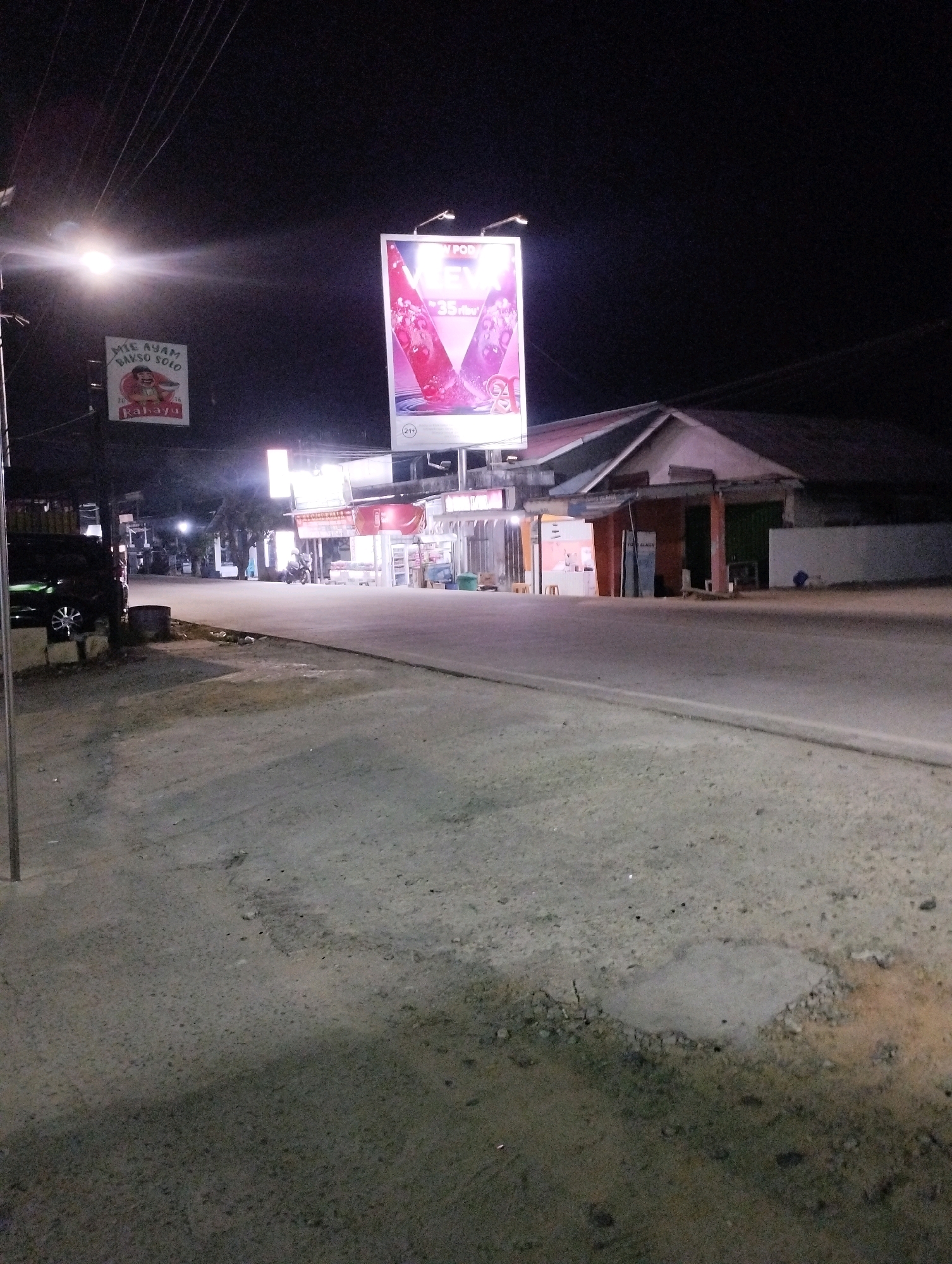 The image depicts a street scene at night in Kuala Samboja, Indonesia. A bright billboard is illuminated and displays a pinkish-red drink with what appears to be ice and fruit. Below this, a smaller sign shows an illustration of a cartoon character with "MIE AYAM BAKSO SOLO" written on it. There are several storefronts with lights on, suggesting they are open for business. A dark-colored car is parked on the left side of the street. The road is paved, with some debris and potholes visible. Power lines stretch across the dark sky, and a street lamp illuminates part of the scene. A few motorbikes are parked in front of the stores, and a person is visible on one of them. The overall impression is of a commercial area at night, with typical street elements.