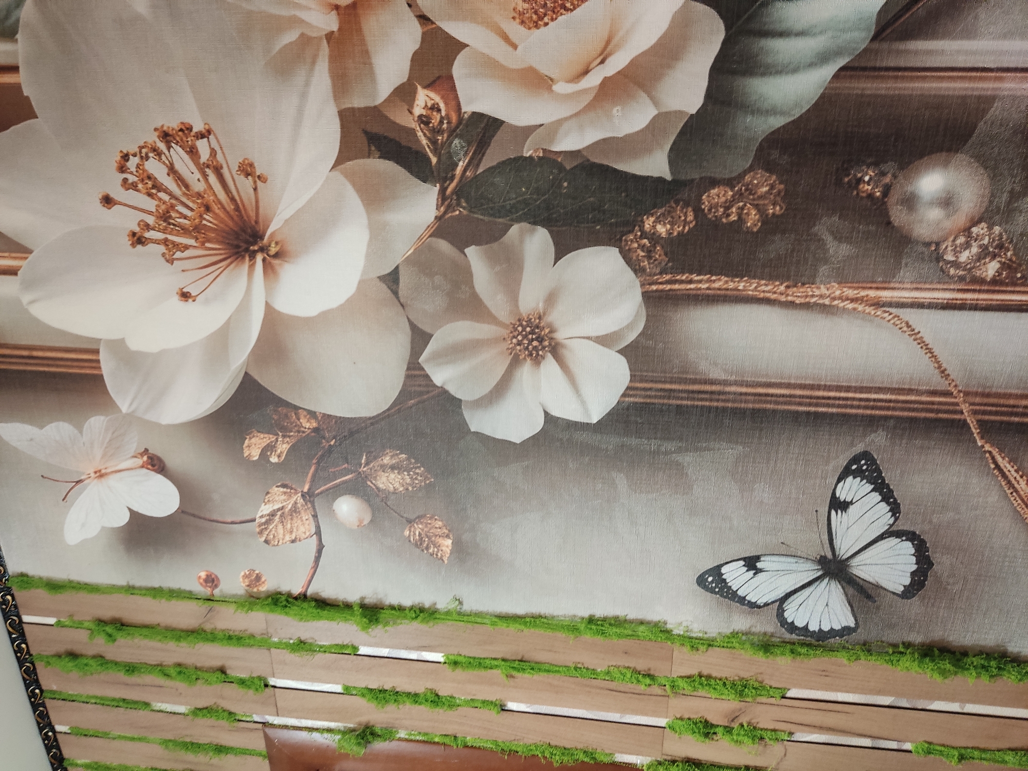 The image shows a wall decorated with a large floral print and moss. The floral print features several large, white flowers with gold accents, as well as two white butterflies. One butterfly is positioned above and to the left of the flowers, while another, larger butterfly with black and white markings, is located in the lower left quadrant of the image. Lush green moss is adhered vertically to a background of wooden slats on the left side of the frame. The overall aesthetic suggests an interior decorative element, possibly a mural or wallpaper, enhanced with natural moss. The location of this scene is in the city of Noginsk, Russia.