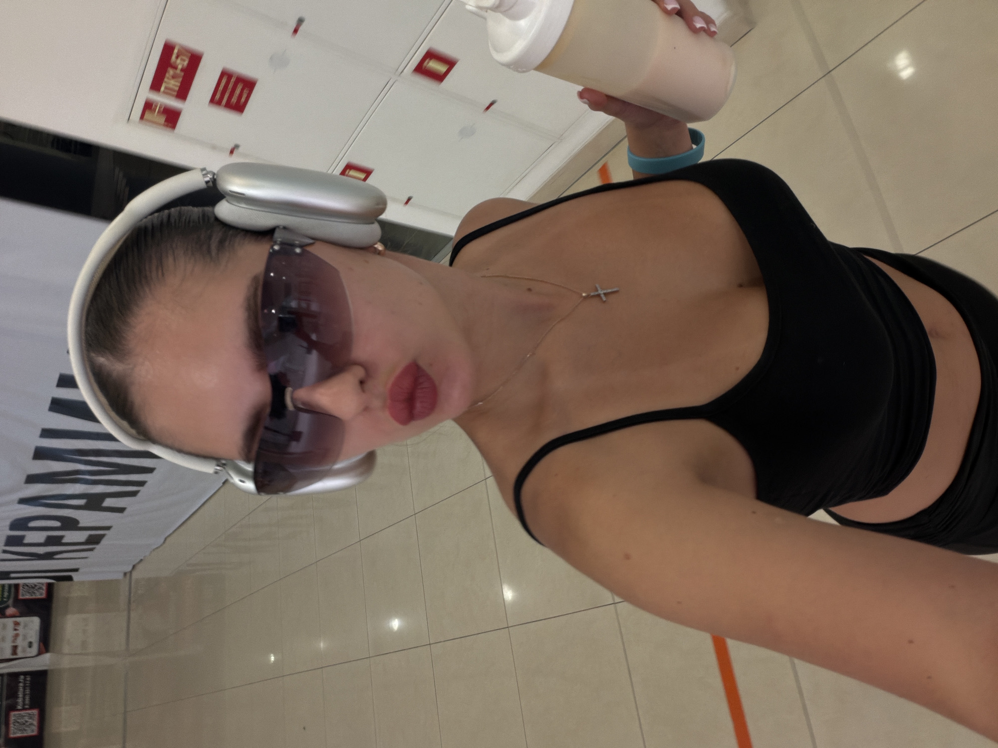 The image is a selfie taken indoors, featuring a young adult female from the chest up, located in Krasnoyarsk, Russia. The female has her hair pulled back and wears large silver over-ear headphones and dark tinted, frameless sunglasses. Her lips are pursed in a reddish-pink lipstick. She wears a black spaghetti-strap crop top, revealing a segment of her midriff, and a silver-colored necklace with a small cross pendant. A light blue silicone wristband is on her left wrist, and she holds a white-capped bottle containing a light pink liquid in her left hand, showcasing fingernails with a French manicure. The background consists of a light-colored tiled floor reflecting overhead lighting. To the left, a large white banner with black Cyrillic text is visible, clearly showing "КЕРАМИКА" (KERAMIKA - Ceramics), alongside a partially visible digital display with QR codes and the text "Kolnatura.ru". To the right, white storage lockers are present, some marked with red rectangular signs, one displaying "ТК1-Б7". The scene suggests a public or commercial interior space, such as a shopping center, exhibition hall, or a fitness facility.