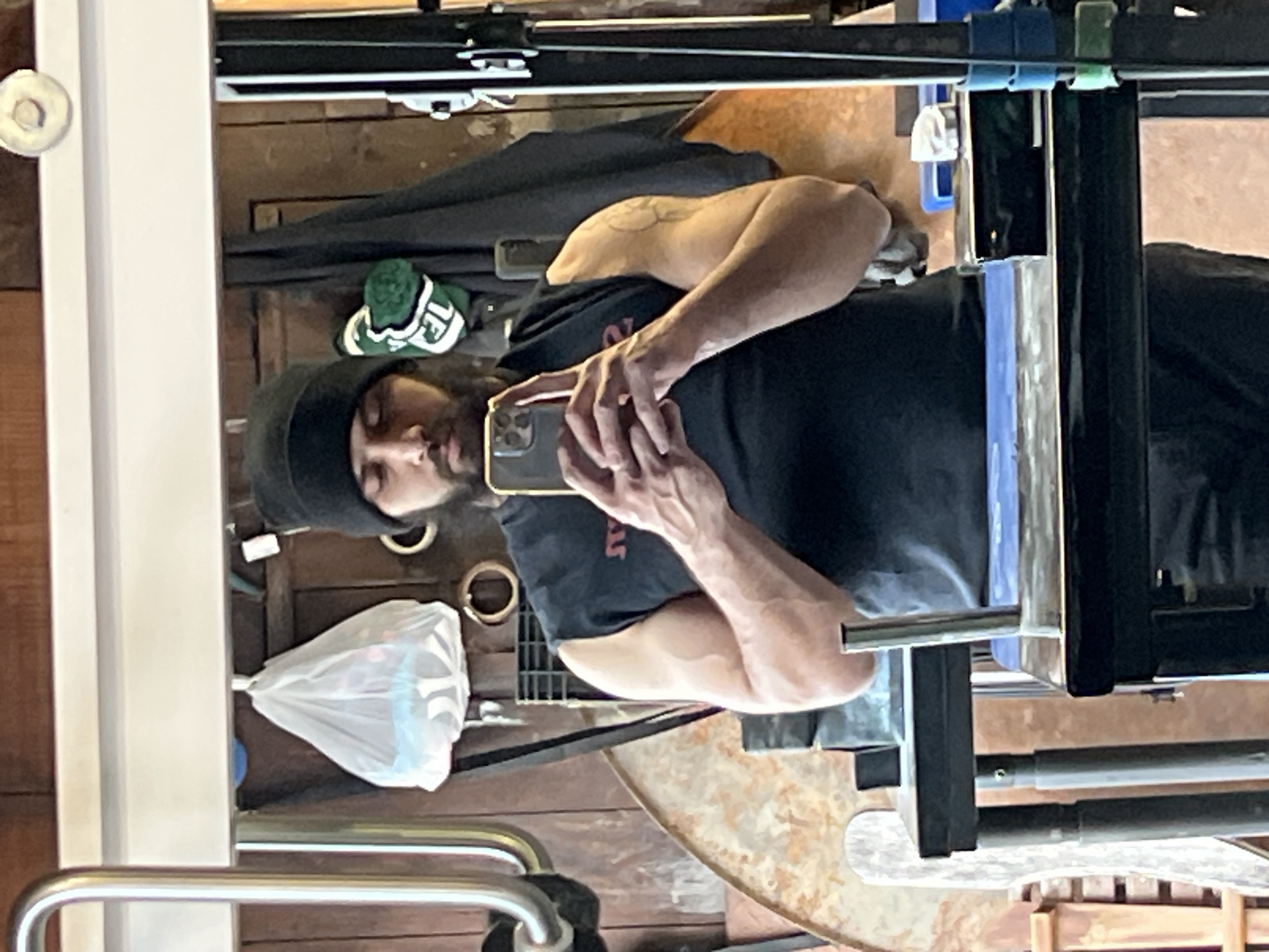 A man is taking a selfie in a mirror. The man is wearing a black beanie and a black t-shirt. The mirror is in a cluttered room. A shopping bag is visible. The image was taken in the City of Newburgh, United States of America.