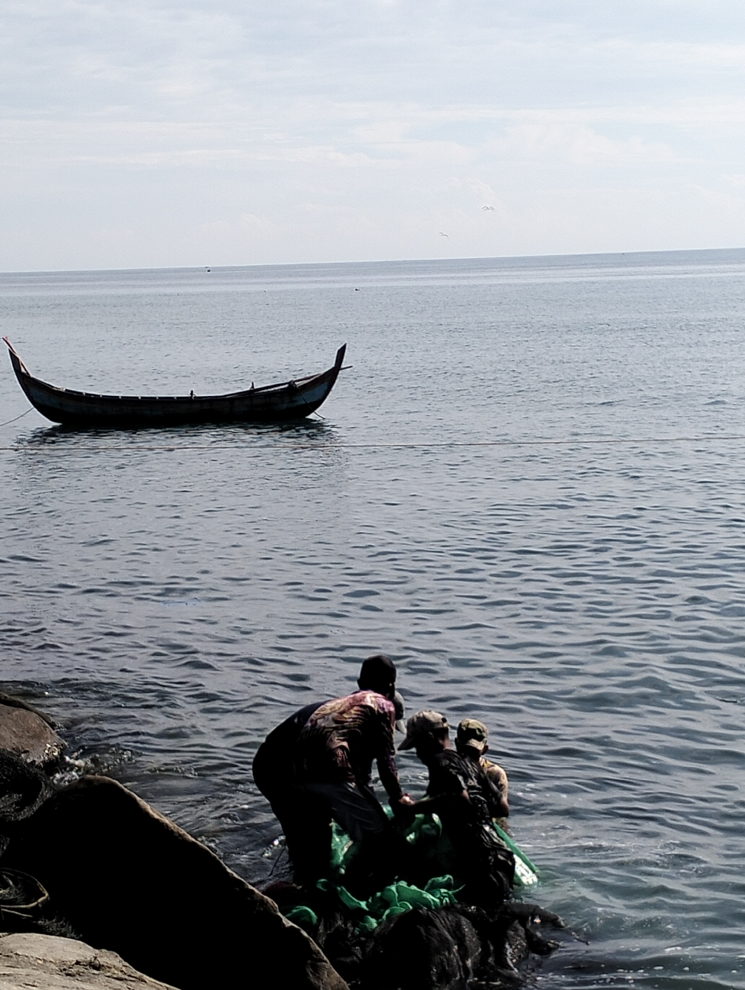 The photograph depicts a coastal scene in Lhokseumawe, Indonesia. A dark-colored, slender boat is anchored offshore. Closer to the shoreline, three individuals appear to be engaged in fishing-related activities; they are standing partially submerged in the water, handling green fishing nets. The immediate foreground consists of rocks and shoreline vegetation. The overall environmental conditions suggest a daytime setting with a cloudy or overcast sky.