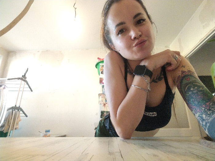 The scene depicts an interior room. No visual identifiers within the image confirm the specified location of Noviy, Russia.
A female subject occupies the lower right and central portion of the frame, leaning forward onto a light-colored wooden surface, possibly a table or countertop. She has dark hair, a circular nose ring, and extensive, colorful tattoos covering her left arm. She wears a black tank top, a black smartwatch on her left wrist, and multiple silver rings and bracelets. Her facial expression shows pursed lips, and she directs her gaze towards the camera.
The background consists of light-colored walls with visible patches of plasterwork or repair. The ceiling above features an exposed light fixture or wiring. To the left, a white collapsible clothes drying rack is present, with a light-colored fabric item draped over it. A small container is visible on the floor beneath the drying rack. A portion of a green plastic bucket is visible on the far right side of the frame.