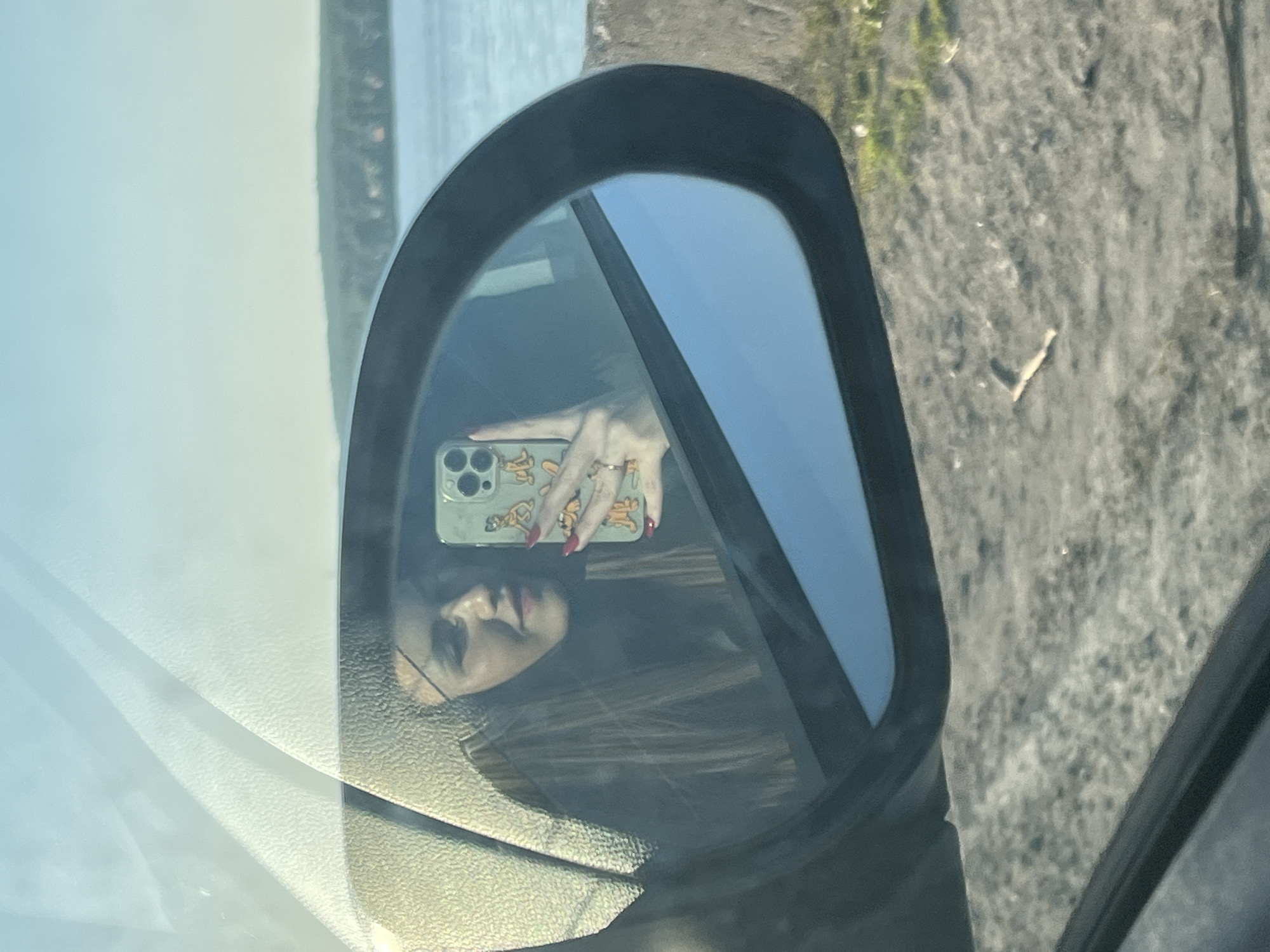 The image shows a reflection in a car's side mirror. The reflection includes a person holding a smartphone with a light green case decorated with cartoon figures. The person's face is partially visible, showing dark hair, eyeglasses, and red lipstick. The hand holding the phone has red nail polish and rings on the fingers. The reflection also shows the car's interior and the outside environment. The background outside the car consists of vegetation and water. Based on the geographic information provided, the photo was taken in the city of Chaikovsky, Russia.