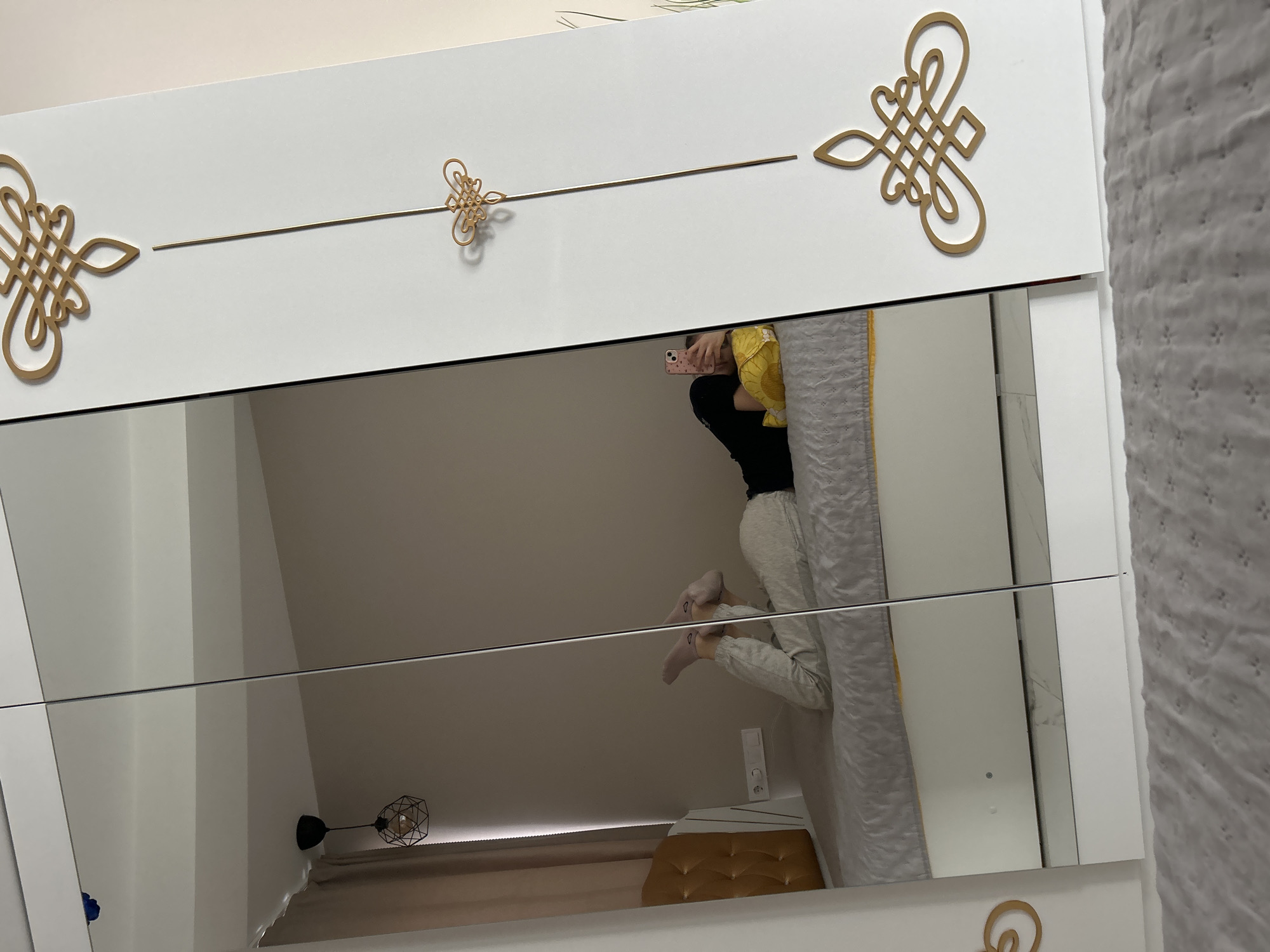 An indoor scene in Sofia, Bulgaria, is captured as a reflection in the mirrored doors of a white wardrobe. A female subject lies supine on a bed with a grey quilted duvet and a yellow accent, wearing light-colored trousers, a black top, and pink socks, with her legs bent and feet elevated. She holds a pink smartphone in her right hand, positioned as if taking the photograph, with her left arm resting on a yellow pillow. Her face is partially obscured by the phone. The reflected background includes a light neutral-colored wall, an upholstered golden-yellow or tan tufted headboard, and a white double electrical outlet. A black geometric caged pendant light fixture hangs from the ceiling, and a beige curtain is partially visible to the left. The wardrobe, which contains the mirrored panels, is white with its rightmost solid panel decorated by three golden ornate knot designs connected by a thin vertical golden rod. The immediate foreground reveals a grey, subtly patterned textile floor covering.