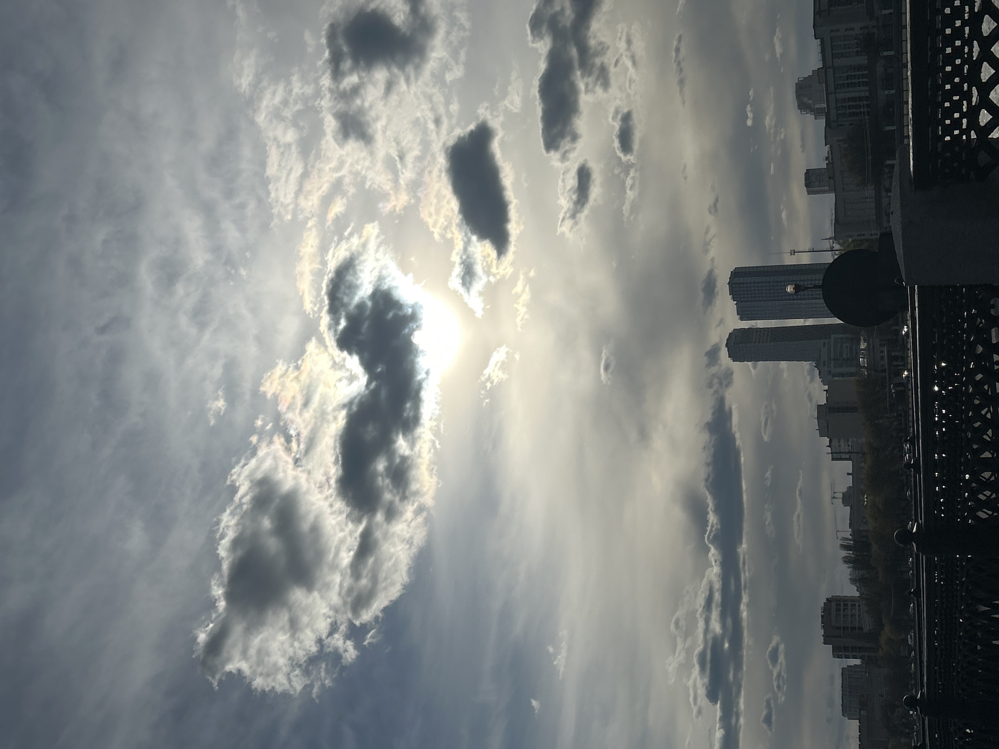 The image captures a dramatic cloudy sky above an urban cityscape, viewed from what appears to be a bridge or elevated platform. The sky, dominating the upper portion, features a dynamic interplay of light and shadow, with large, dark clouds contrasting against bright, illuminated wisps. The sun, partially obscured by a dense cloud in the upper left, casts a brilliant glare and creates iridescent, rainbow-like edges on the surrounding clouds.
Below the expansive sky, a silhouette of a city skyline stretches across the horizon. Several tall, modern buildings, possibly residential or commercial high-rises, are visible, their dark forms indistinct against the brighter background. On the right side of the frame, a dark, ornate metal railing, indicative of a bridge or balcony, provides a strong structural element. A round, dark object, possibly a lamppost or a decorative fixture, is also visible near the railing.
The scene depicts daytime conditions, characterized by bright, albeit obscured, sunlight filtering through the clouds. The weather appears to be partly cloudy, creating a high-contrast and atmospheric view. No people or discernable text are visible in the image. The setting is an urban environment, identified as Moscow, Russia, offering a glimpse of the city's architecture under a dynamic sky.