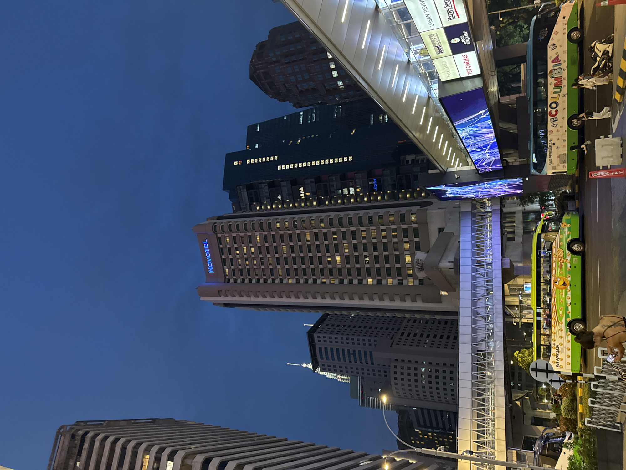 This is an exterior, ground-level, night-time shot, appearing to be in the city of Kuala Lumpur, Malaysia. Several tall buildings of modern design are present, including a "NOVOTEL". Also present are the brands and signs such as "Harvey Norman", "TGV Cinemas", "foodrepublic", "PARKSON". A "COLUMBIA" labeled bus is seen, along with pedestrians nearby. Another lime green bus with a smiling face is present. An elevated, enclosed walkway is suspended between buildings. The sky is a dark blue, indicating that it is night. A sign reading "COVALET" is also seen. The image is taken from below, looking upwards towards the buildings.