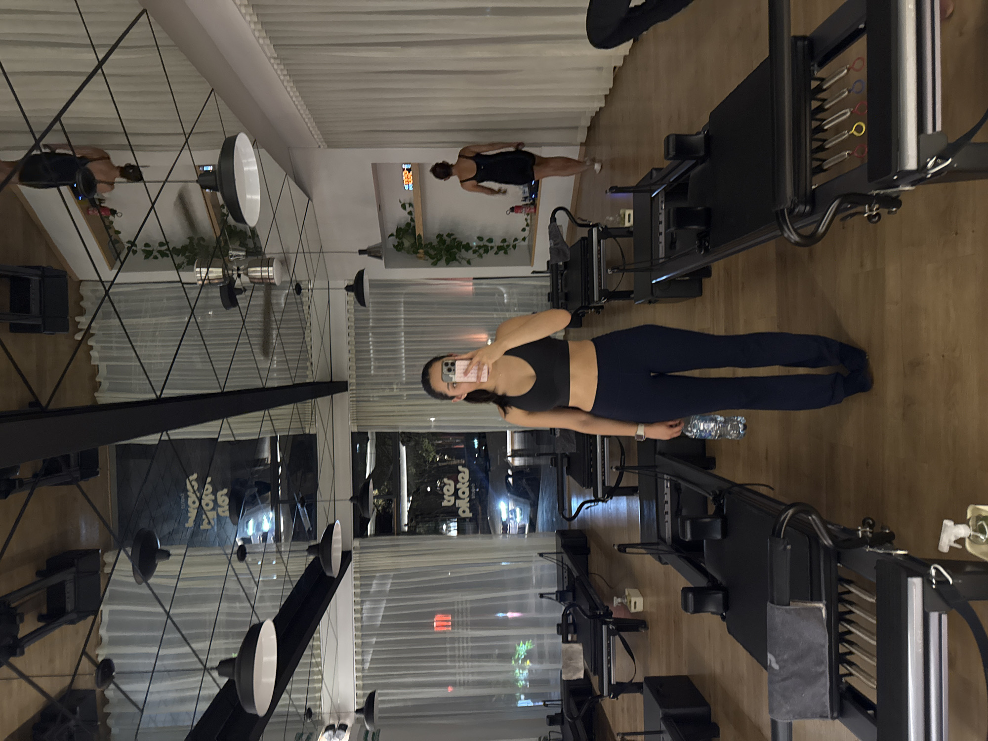 The image depicts the interior of a Pilates studio, likely in Puebla, Mexico, given the reflection of a sign reading "tres pilates ESTUDIO". The studio has wooden flooring and is equipped with Pilates reformer machines. A woman in a black sports bra and blue leggings is holding a smartphone, presumably taking a selfie in front of a reformer. Another person in a black dress is visible in the background, standing near another reformer. There are mirrors and hanging lights on the ceiling. A water bottle and a spray bottle are placed near the reformers. Through the window, an urban environment is visible with streetlights and signage.