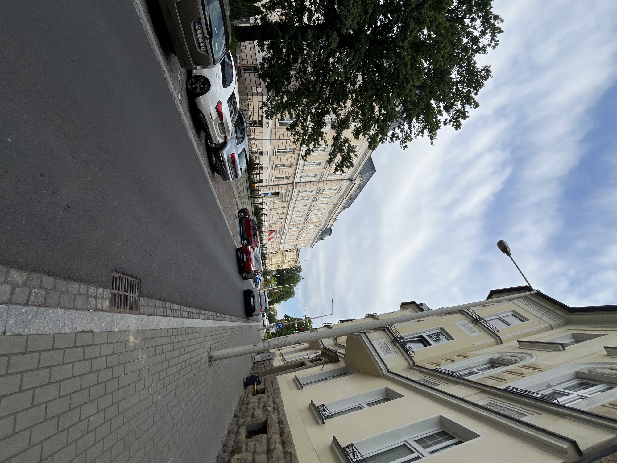 The image shows a street scene in a city, with buildings on both sides of the road and cars parked along the curb. The buildings appear to be old, with architectural details such as ornate facades and balconies. The street is paved with asphalt, and there is a sidewalk made of cobblestones. A large tree is visible on the left side of the image, and the sky is cloudy. Several cars are parked on the street, including a white sedan, a silver SUV, and a dark-colored hatchback. A red hatchback is driving down the street, and a dark gray hatchback is also visible in the distance. There are no people clearly visible in the image. The overall impression is one of a quiet, residential street in an older part of the city.