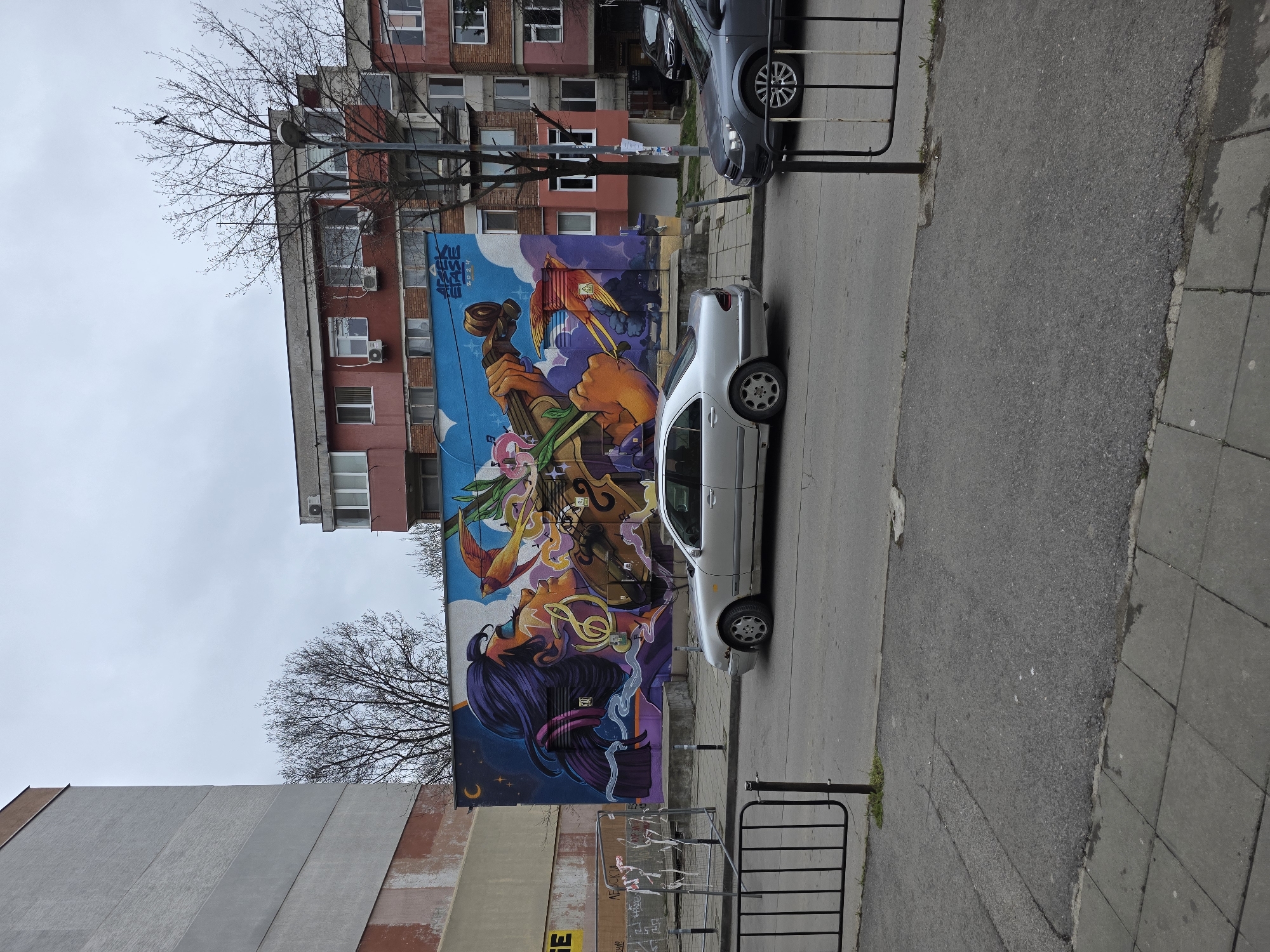 The image depicts a street scene in Sofia, Bulgaria. A large, colorful mural covers the side of a building, depicting a woman with flowing purple hair, musical notes, and a bird. A silver car is parked on the street in front of the mural. A dark gray car is parked ahead. A black metal fence stands on the sidewalk. Another building is seen behind the first. The sky is overcast.