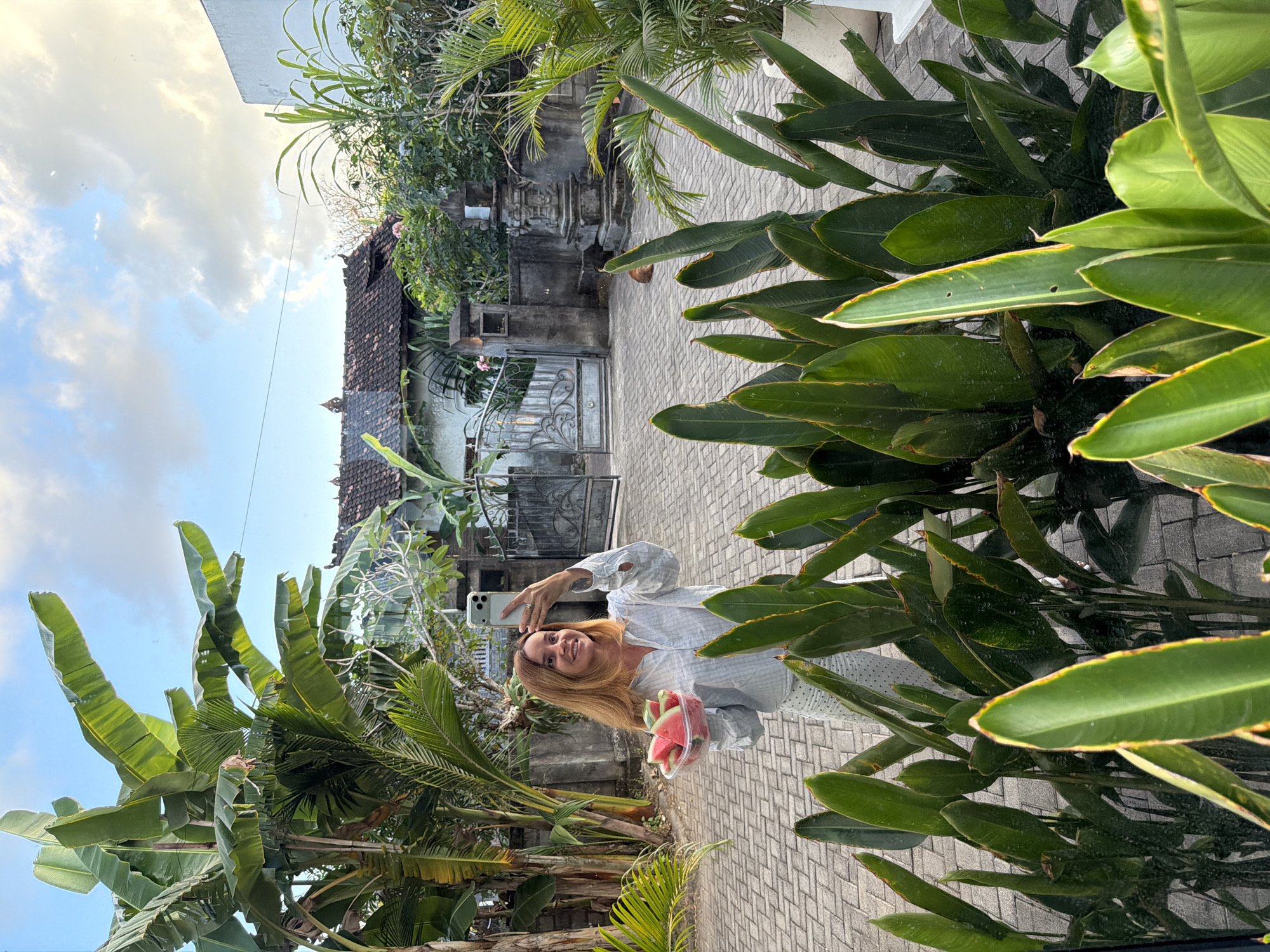 In Kuta Selatan, Indonesia, a woman with auburn hair stands on a paved courtyard, taking a selfie with her smartphone while holding a clear plastic container filled with watermelon slices. She is wearing a white striped shirt and patterned white pants. Behind her, a grey wrought-iron gate leads to a traditional Indonesian house with a tiled roof. Lush green foliage, including banana trees and other tropical plants, surrounds the scene, framing the courtyard and the residential building. The sky above is partly cloudy with a light blue hue.