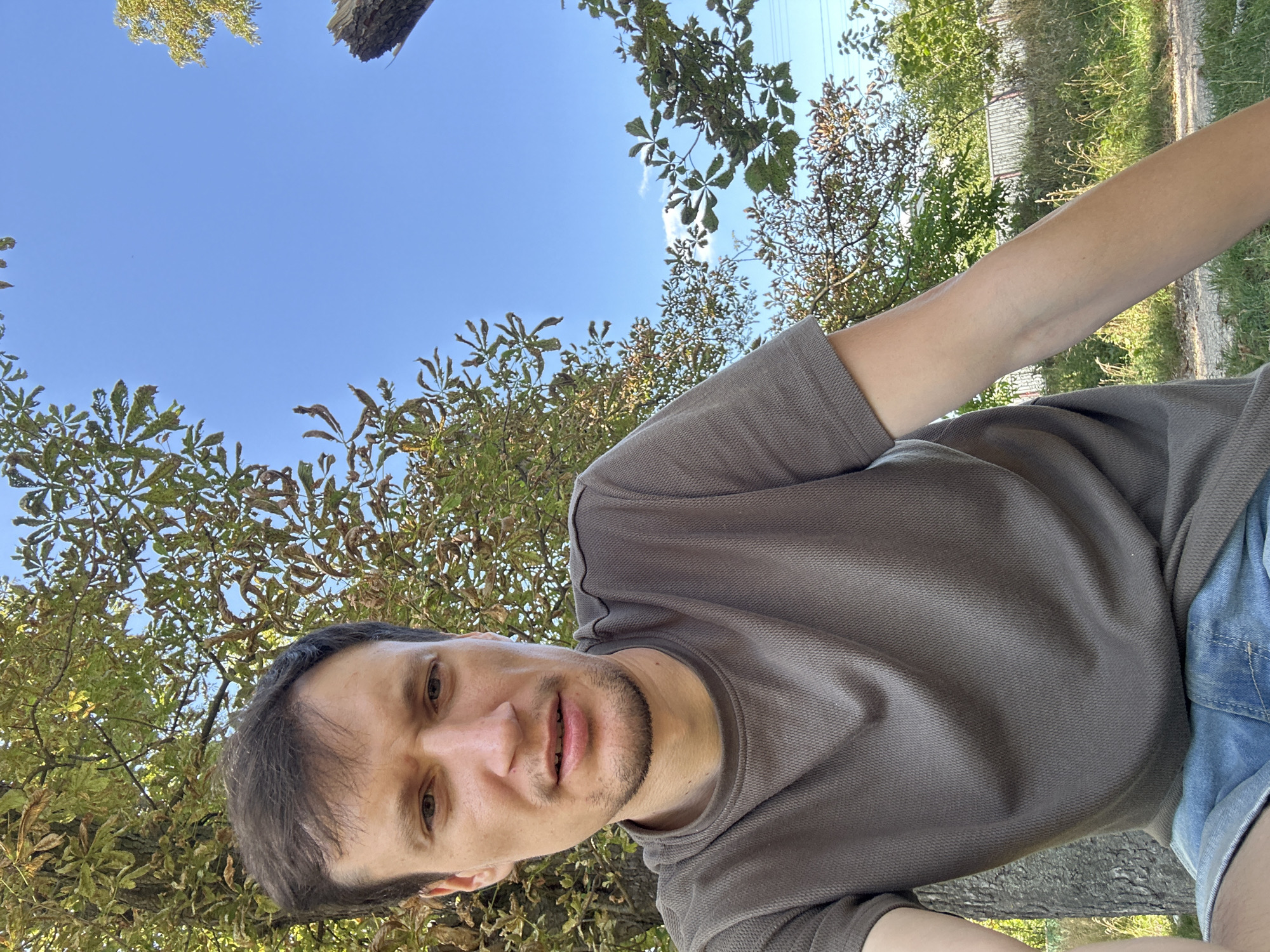 A man is outdoors, with a brown textured t-shirt and blue denim shorts. He has brown hair and a light complexion. He is looking at the camera. He is sitting in front of a tree with green and brown leaves. The sky is blue with a few white clouds. The background includes more trees and a fence. The location is Makiv, Ukraine.