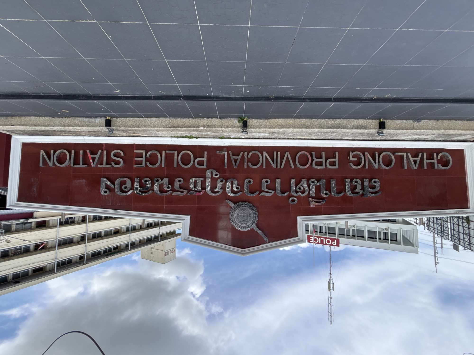The image shows a sign for the Chalong Provincial Police Station in Chalong, Thailand. The sign is dark red with silver lettering. The top line of text is in Thai script, and the bottom line reads "CHALONG PROVINCIAL POLICE STATION" in English. Above the Thai text is a circular emblem with a sword crossed behind it. In the background, two police buildings are visible. One is white with "POLICE" written in red letters, and the other is a multi-story beige building with many windows. The sky is cloudy. The foreground is a tiled walkway.