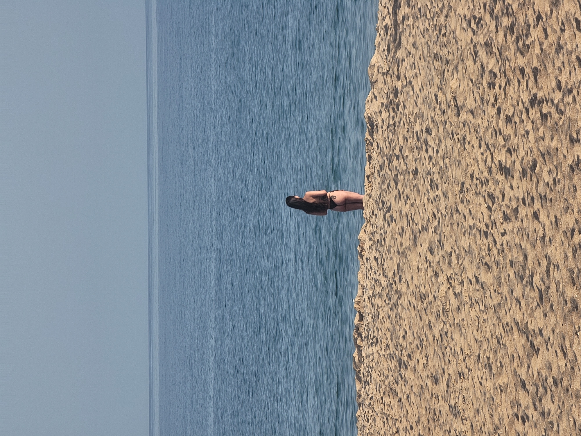 A woman stands on a sandy beach, facing the ocean. She has long dark hair and is wearing a black bikini. The water is a deep blue, with gentle waves. The sky above is a pale blue. The sand is a light brown color and appears to be undisturbed except for footprints. The scene is peaceful and serene. The location is Quarteira, Portugal.