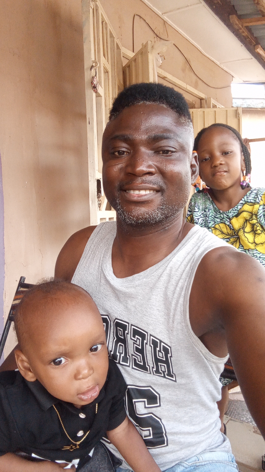 The image shows a man in Sabon Tasha, Nigeria, holding a baby and with a young girl standing behind him. The man is wearing a gray tank top with black lettering and has graying facial hair. The baby is wearing a black shirt and gold necklace. The girl has braids and is wearing a colorful floral print dress. The location appears to be outside a building with a light-colored wall and decorative metalwork.