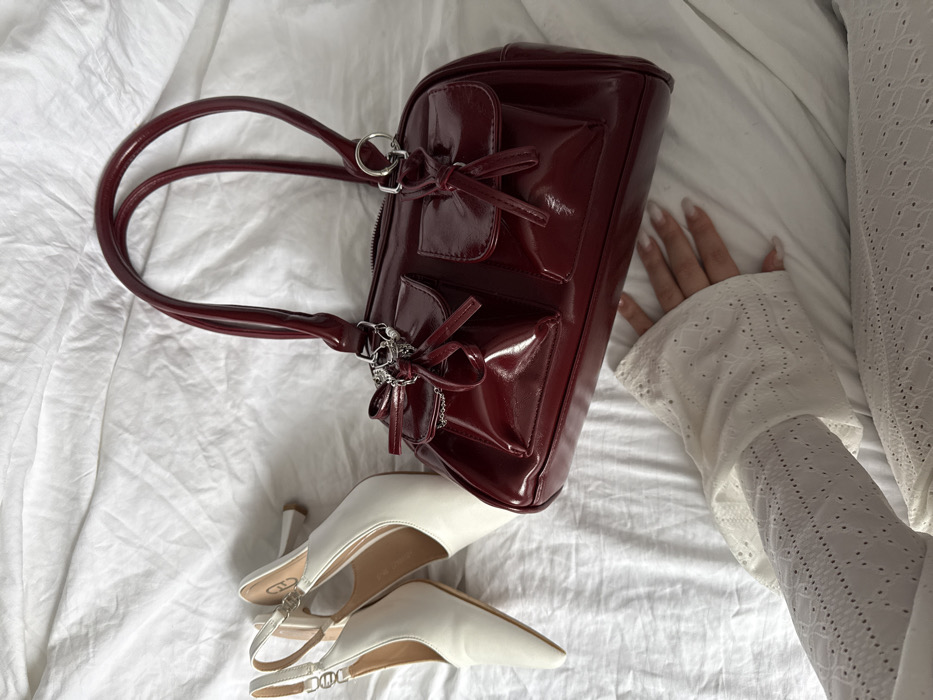 The image displays an indoor scene featuring personal items arranged on a white, wrinkled textile surface. A dark red, high-gloss handbag is centrally positioned, characterized by two rounded top handles, a zipper closure, and two front flap pockets each secured with a decorative tie. Silver-colored metal rings and a short chain detail are attached near the handbag's handles. To the upper left, a pair of white pointed-toe slingback shoes with block heels are present; one shoe displays a light-colored logo on its inner strap. In the lower frame, a human arm and hand are visible, resting on the white fabric. The arm is covered by a white long-sleeved garment featuring an intricate eyelet pattern. The visible hand has manicured nails with a light, opaque polish. The environment is domestic and interior; no specific external landmarks or architectural features are present to determine a precise location within Krasnodar, Russia.