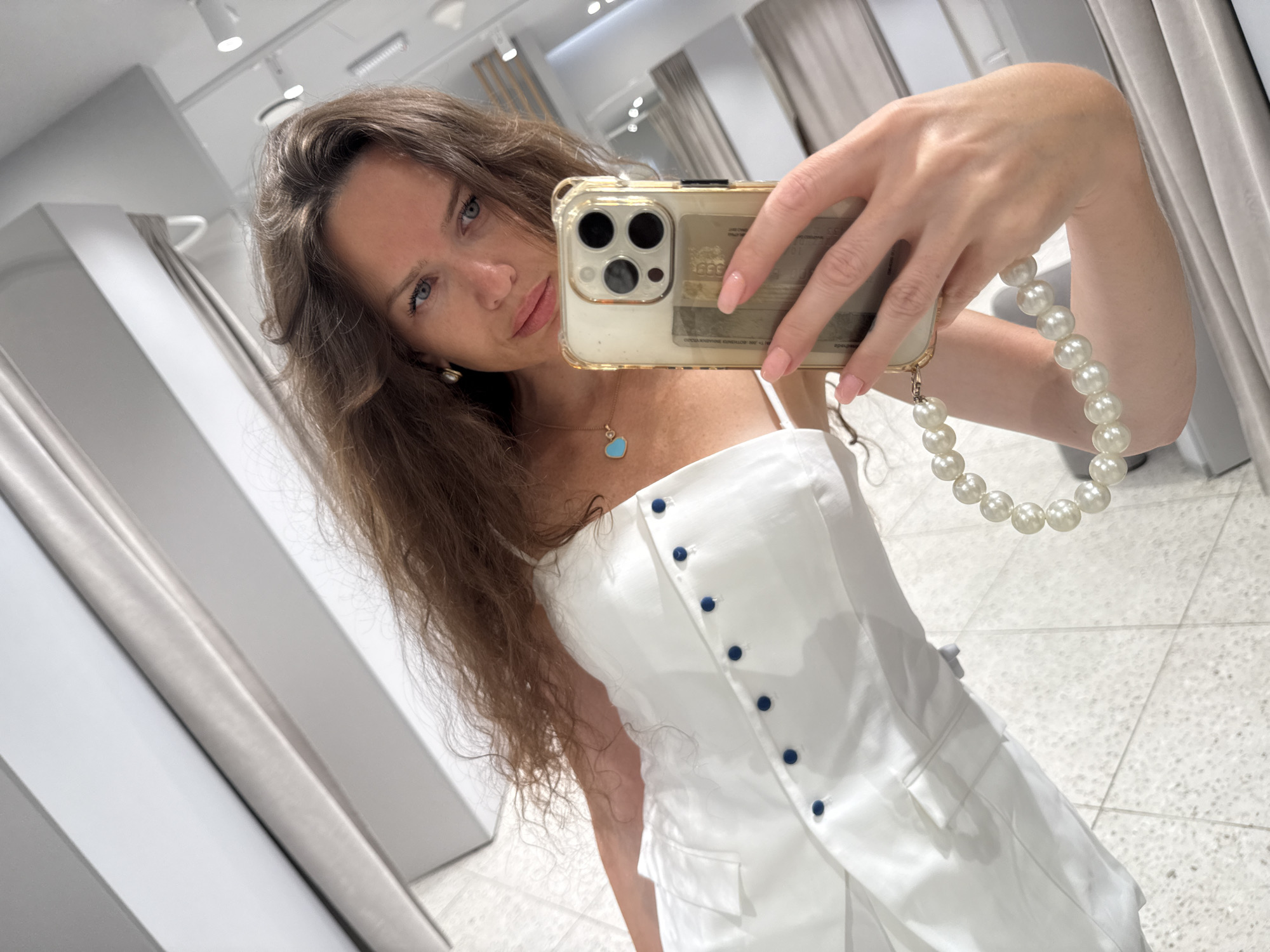 A woman with long, brown, wavy hair is taking a selfie in what appears to be a fitting room or a closet. She is wearing a white garment with blue buttons and a blue heart-shaped pendant necklace with a turquoise stone. She is holding a gold-colored smartphone with a clear case that has a cardholder attachment, and a pearl strap is attached to the phone. Her nails are painted pink. The background is a mix of reflective surfaces and what appear to be clothes hanging on racks, suggesting a retail environment. The lighting is bright, and the floor has a textured, tiled pattern. There are no immediate indicators of the specific city or location beyond it being indoors.