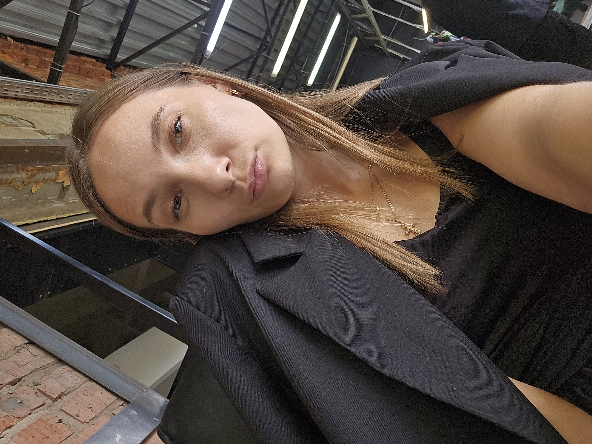 The media features a close-up selfie of a young woman, likely in her 20s or early 30s, with light brown hair and brown eyes. She looks directly at the camera with a calm, slightly pensive expression and gently parted lips. Her head is tilted, appearing to rest on her left shoulder, which is covered by a black blazer. She wears a black top underneath and a delicate gold necklace with a small charm around her neck.
The background suggests an indoor setting with an industrial or urban-chic aesthetic. Above her, exposed metal beams and a ceiling that appears to be made of corrugated metal are visible, illuminated by several bright fluorescent tube lights. To her left, an exposed brick wall adds to the industrial feel, along with distressed wall textures and a metal railing in the lower left foreground. The angling of the photo creates a dynamic and intimate perspective. The scene is located in Chaikovsky, Russia. There is no visible text in the image.