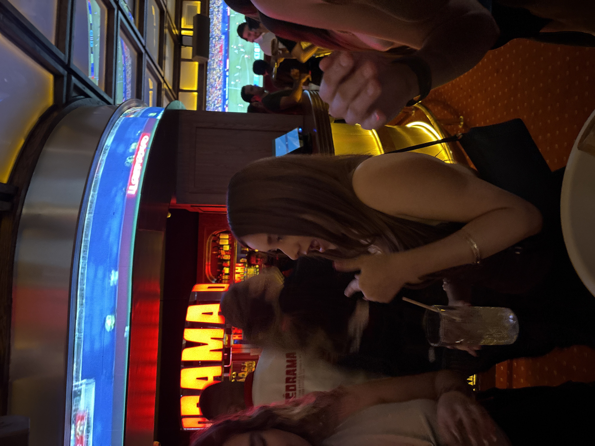 The image depicts a lively interior scene, likely a bar or club, in San Pedro Garza García, Mexico. In the foreground, a young woman with long brown hair is captured mid-conversation, gesturing with her right hand. She is wearing a dark top and a bracelet. To her left, another woman's shoulder and arm are visible, wearing a light-colored top. In the background, a man wearing a white jacket with "EDRAMA" written on the back is visible. Several other individuals are present, some blurred due to motion. The environment is illuminated by neon lights, with a large screen displaying a sports game, possibly soccer, in the upper portion of the frame. Another screen with blue light is visible to the left, and a bar area with bottles is in the background. The floor is covered with a red carpet with a patterned design. The overall atmosphere suggests a social gathering or entertainment venue.