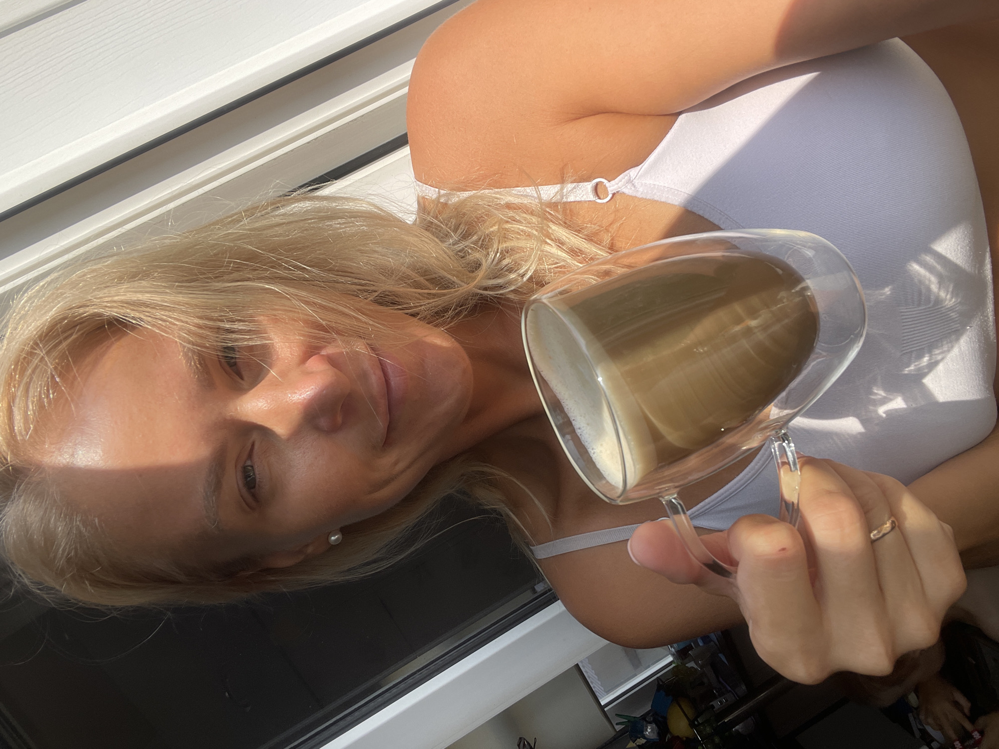A blonde woman holds a clear, double-walled glass mug filled with a light brown, frothy beverage. She is wearing a white tank top. The woman is looking towards the camera with a slight smile. Sunlight casts a shadow across her face and hair. The background appears to be indoors, with a white door or window frame visible on the right and a dark area on the left that might be a window looking outside. There is also a hint of furniture or objects in the far left background. The overall scene is brightly lit by natural light, suggesting it is daytime.