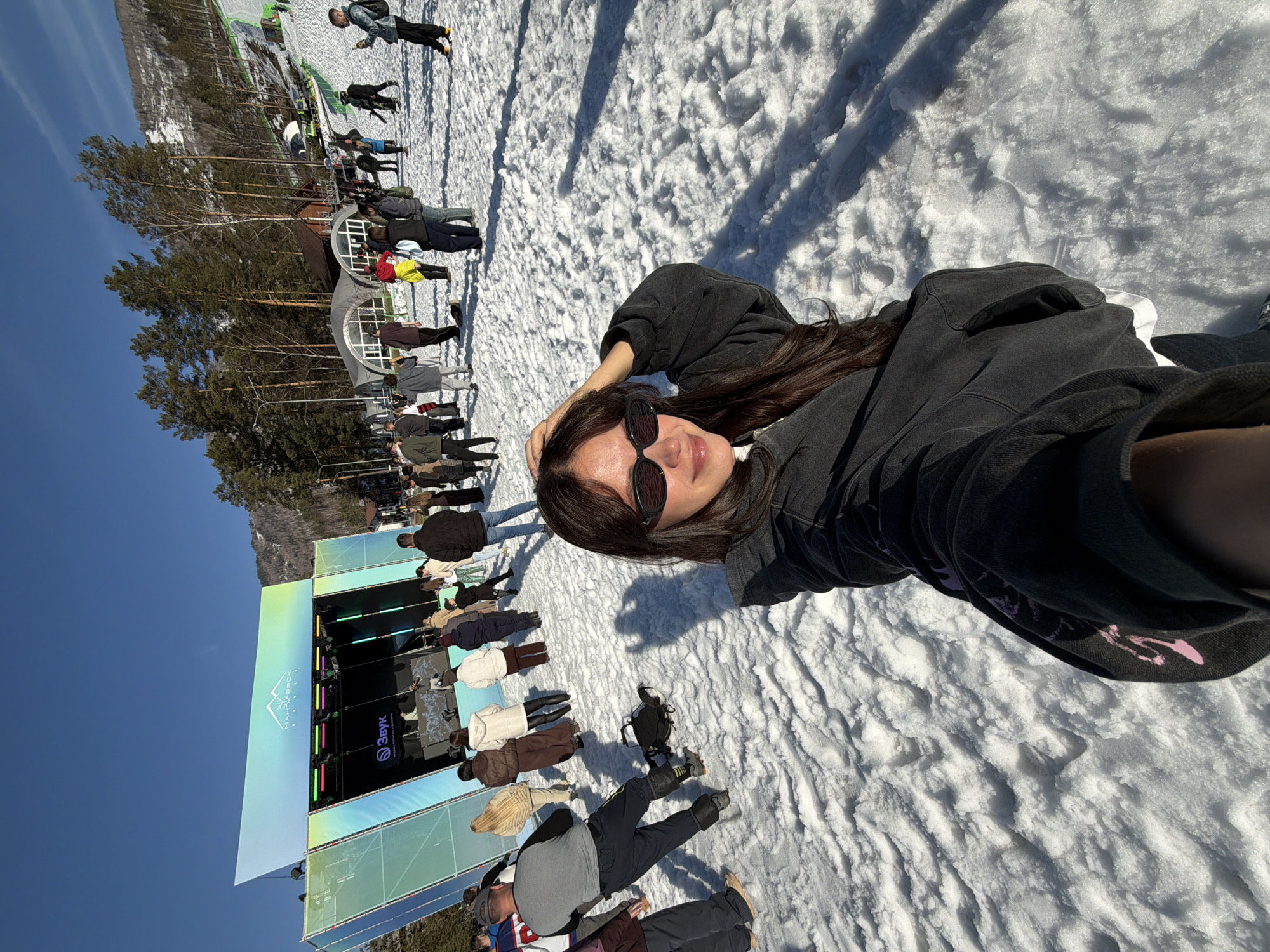 A woman with long dark hair and sunglasses lies on snow, taking a selfie with a crowd of people and a stage in the background. The stage features a large screen and the words "Malinovka Fest" in Russian. The location appears to be Озёрное, Russia, as the festival is known to take place there. The sky is clear and blue.