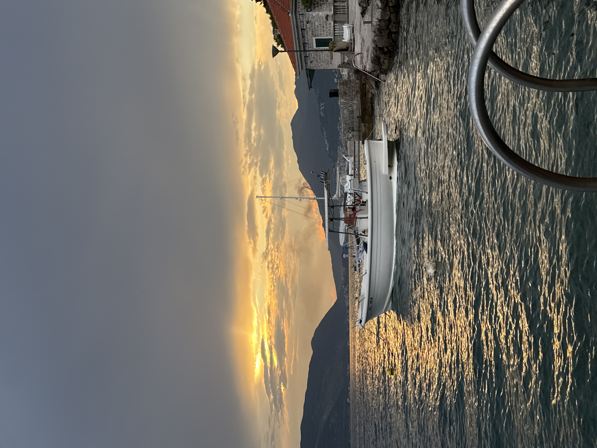 A boat is docked next to a stone building with a red-tiled roof. The boat is white with a mast and rigging. The water is rippled and reflects the golden light of the setting sun. Mountains rise in the background, silhouetted against a sky filled with dramatic clouds. The scene suggests a peaceful evening by the water, perhaps in a coastal town. The overall mood is serene and picturesque. The time of day is clearly sunset, indicated by the warm, low-angled light and the colors in the sky. The weather appears calm, with the water smooth and the clouds illuminated. The architecture of the building suggests a European setting, possibly Mediterranean. The boat appears to be a sailboat or a motor yacht. There is some visible text on the boat, "ZL 154."