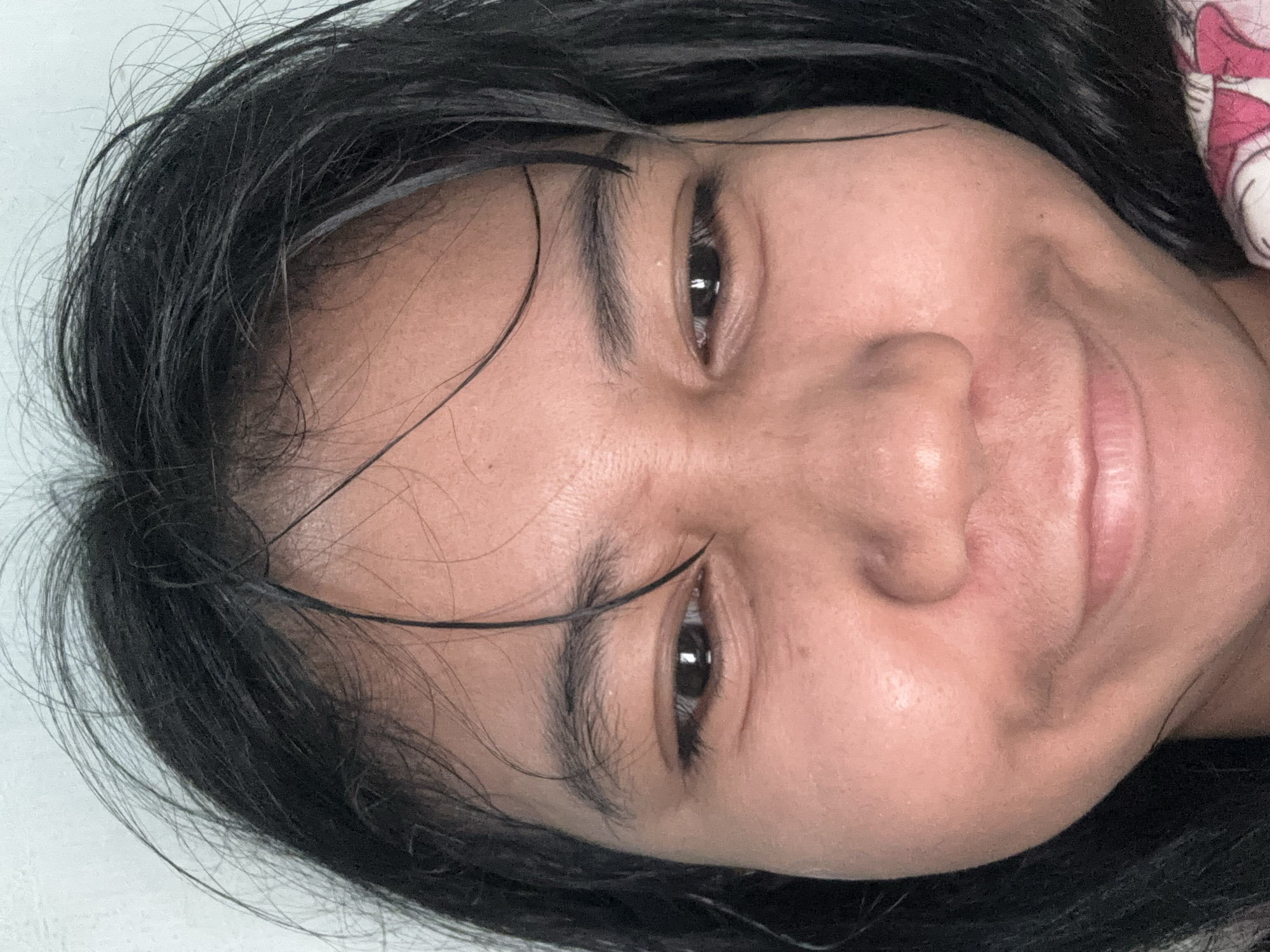 This is a close-up, waist-up photograph of a young East Asian female. Her skin is a light tan, and her dark hair is wet and messy, falling around her face. She is looking downwards with a slight smile, her eyes are dark brown and her eyebrows are thin and dark. The background is a plain white wall, suggesting an indoor setting. The lighting is even and appears to be natural light coming from the front. There is no indication of the specific location within North Jakarta, Indonesia in the image itself.