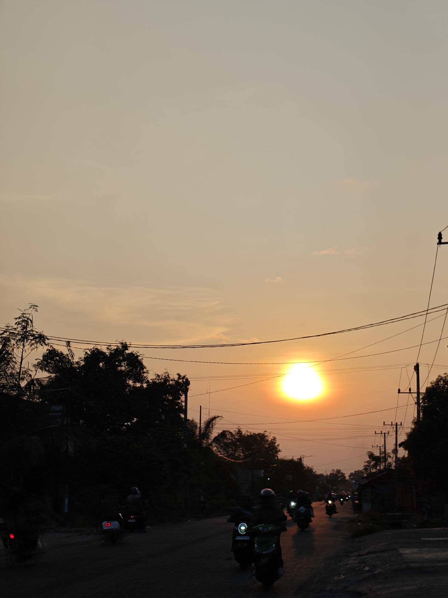 The image depicts a street scene in Blangcot Tunong, Indonesia, at what appears to be sunset. A large, intense, orange-yellow sun is prominent in the upper-middle right quadrant of the frame, casting a warm hue across the sky, which transitions from bright orange near the sun to a muted beige-orange at the top. The sky contains minimal, faint cloud cover.
The lower two-thirds of the image are significantly darker, primarily featuring silhouetted elements against the bright sky. A road, potentially asphalt or compacted dirt, extends from the foreground into the distance, appearing somewhat reflective in certain areas, possibly due to moisture or light conditions. Numerous motorcycles or scooters are traveling on this road, mostly visible as dark silhouettes. Several vehicles have illuminated headlights or taillights (e.g., a red taillight on a scooter on the far left, a white headlight on a scooter in the mid-ground, and multiple smaller lights further down the road). Riders, also in silhouette, are discernible on the motorcycles; some appear to be wearing helmets.
Dense vegetation, including trees and bushes, flanks both sides of the road, appearing as dark, undifferentiated masses against the lighter sky. Overhead utility poles with a network of electrical wires are visible, particularly on the right side of the frame, also rendered in silhouette. A few indistinct structures or buildings are partially visible behind the trees on the sides of the road. No specific interactions between individuals are discernible; the primary action is the movement of traffic along the road.