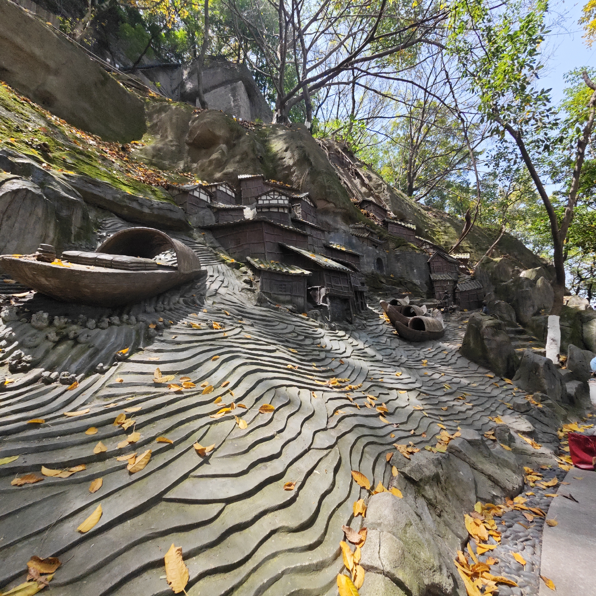 The image depicts a hillside in Yuzhong District, Chongqing, China. The scene showcases a detailed miniature representation of traditional riverside housing. Small, dark-wood houses are constructed on the slope, with two boat sculptures situated among the houses. The "water" is represented by undulating concrete, covered in scattered yellow leaves. Mature trees surround the hillside, and the sky is visible through their branches. A pedestrian path is on the right side of the image.