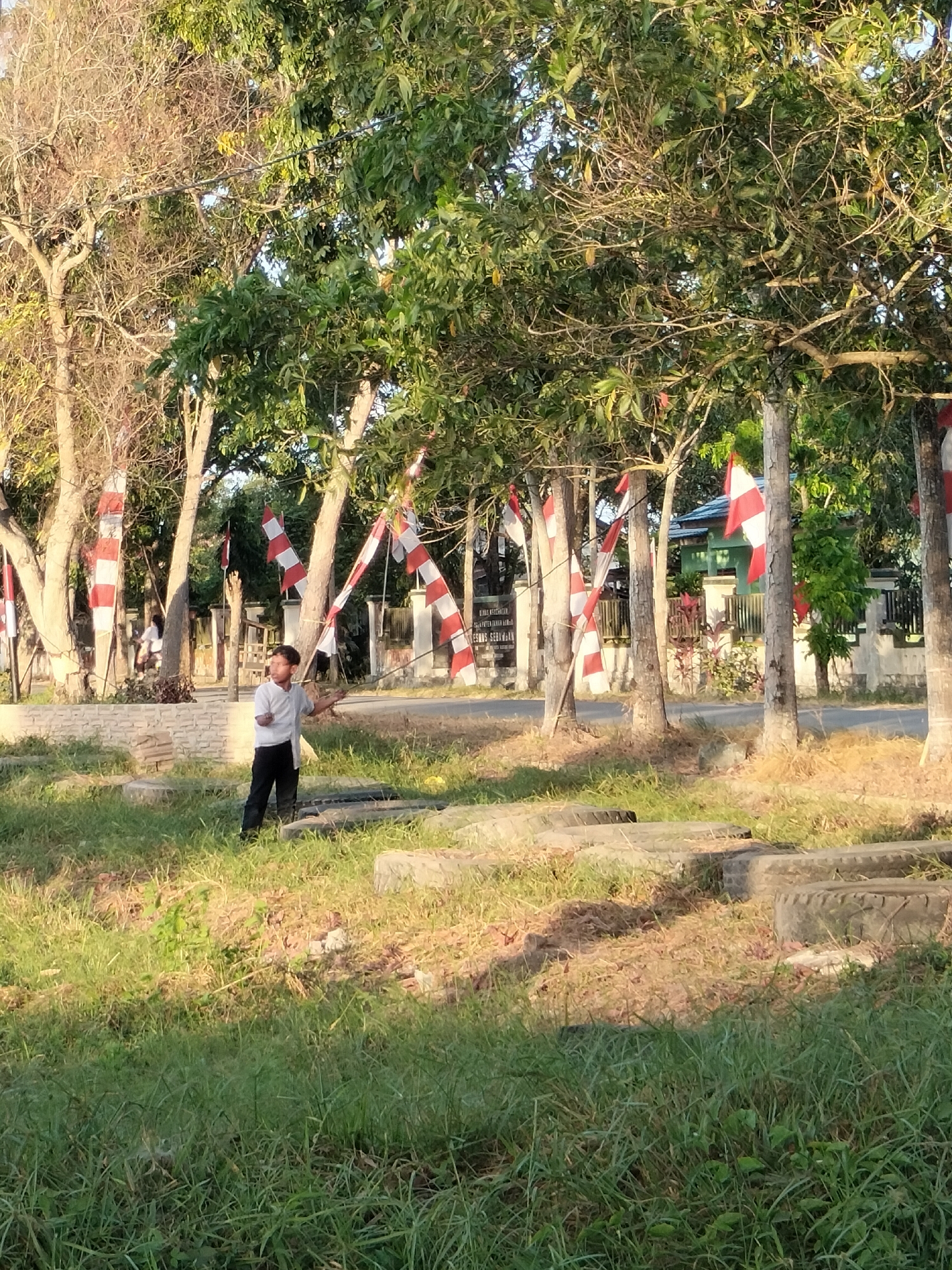 In the city of Karang Indah, Indonesia, a young male is seen standing in a grassy area next to a road. He is wearing a light-colored t-shirt and dark pants, and appears to be holding a long, thin object. In the background, trees are decorated with red and white striped flags. A building with a green roof and a white fence is visible behind the trees. The ground is covered in grass and some patches of dry leaves, with several circular concrete structures or tire bases scattered throughout the foreground. The lighting suggests it is either early morning or late afternoon.