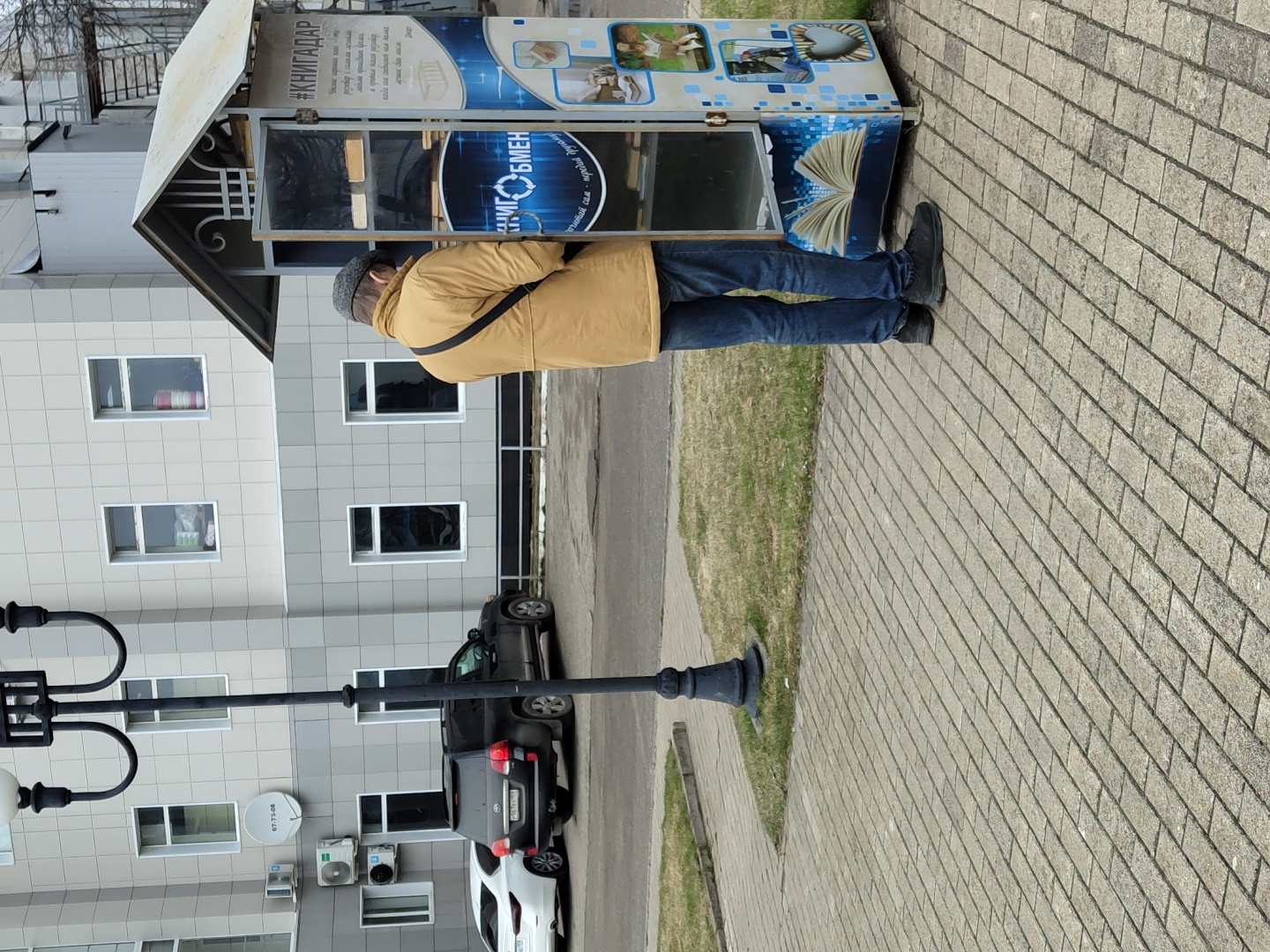 An outdoor, slightly rotated, eye-level shot captures a person standing in front of a book exchange kiosk on a paved sidewalk in Chaikovsky, Russia. The individual, seen from behind, wears a mustard yellow jacket, blue jeans, dark shoes, and a dark beanie or cap, with a dark bag strap visible across their back. They are facing a blue and white book exchange kiosk, which features images of open books and animals, and has a small, light-colored roof.
Visible Russian text on the kiosk includes "#КНИГАДАР" (Book Gift), "КНИГОБМЕН" (Book Exchange), "почитай сам" (read it yourself), and "передай другому" (pass it on to another), along with the name "Декарт" (Descartes). Fragments like "розевер с силами има" and "в правом никой разову" are also visible but less clear.
To the left of the person is a multi-story building with a modern grey tiled facade and multiple rectangular windows. A satellite dish and air conditioning units are mounted on its side, with the number "67-73-08" visible near them. In the foreground and midground, a black street lamp with two lights stands prominently. Several cars, including a black SUV and a white sedan, are parked near the building. The ground is covered with light grey paving stones, and a strip of green grass separates the sidewalk from a road. The lighting suggests daytime under an overcast sky or soft sunlight.