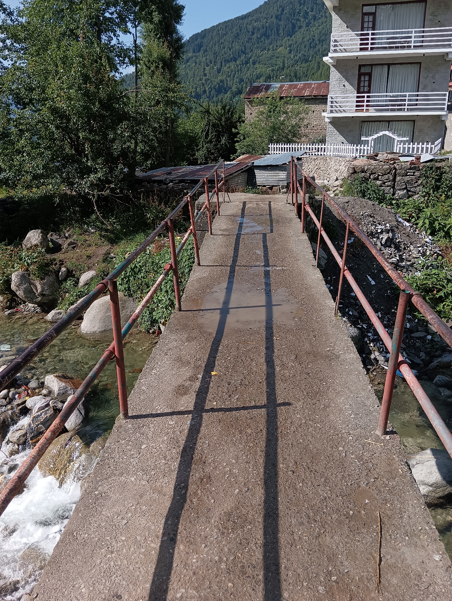 A concrete bridge spans a narrow, rocky stream in a mountainous area. The bridge has rusty metal railings on either side, painted red and green. The stream flows rapidly with white foam, and rocks are visible in the water and along the banks. Lush green trees and foliage grow on the left bank. On the right bank, there is a pile of debris, including rocks and what appears to be refuse. In the background, a multi-story building with balconies and white railings stands to the right, constructed of light-colored stone. To the left of the building, there is an older stone structure with a rusted metal roof. A white picket fence runs along the edge of the property near the buildings. The scene is set against a backdrop of heavily forested mountains under a clear blue sky. This image was taken in Shanag, India.
