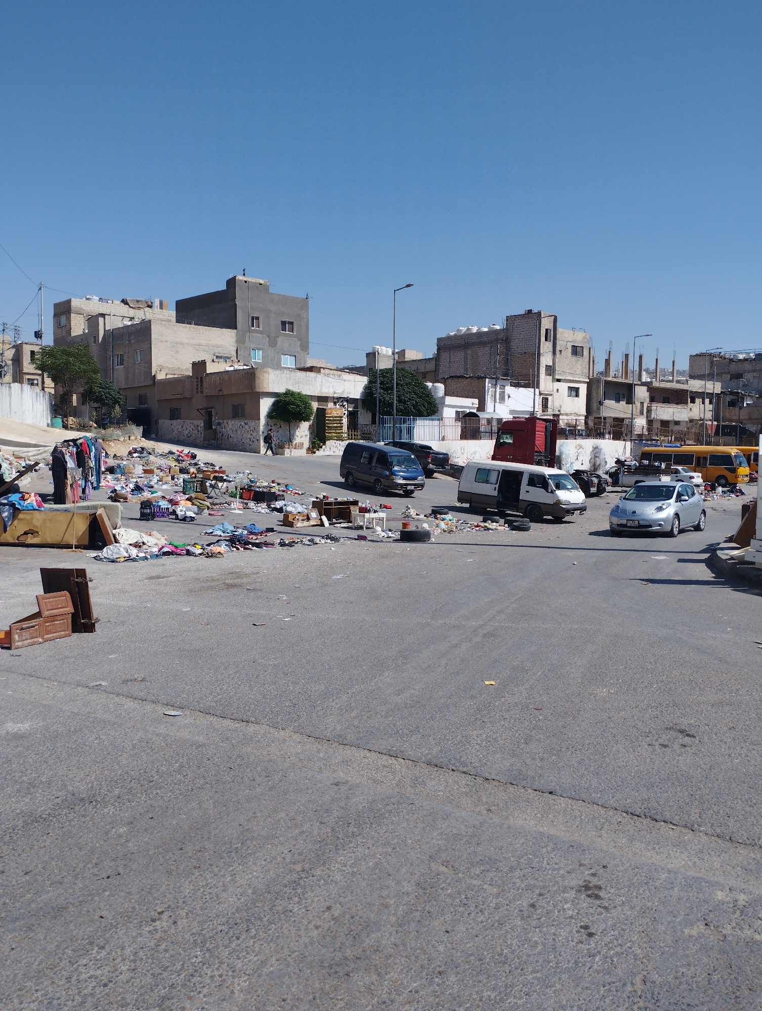 The image depicts an outdoor market scene in the city of Baqa'a camp, Jordan. A vast amount of assorted items, predominantly clothing and household goods, are spread across the asphalt ground, suggesting a flea market or informal trade area. Several vehicles are present, including a silver sedan, a white van, a dark van, and a yellow bus, indicating it is a public thoroughfare or parking lot. In the background, there are multi-story residential buildings, typical of a camp environment, with some showing signs of construction or disrepair. The sky is clear and blue, suggesting a sunny day. The overall scene is chaotic and cluttered, with visible debris and discarded materials scattered throughout the area. There are a few individuals present, some of whom appear to be interacting with the merchandise, possibly as vendors or shoppers.