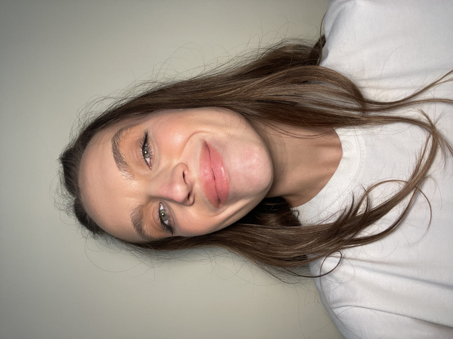 A woman with light skin, brown hair, and green eyes smiles at the camera. She has full lips and light makeup. Her hair is long and loose, falling around her face and shoulders. She wears a plain white t-shirt. The background is a pale gray or off-white color. The image is rotated slightly counter-clockwise.