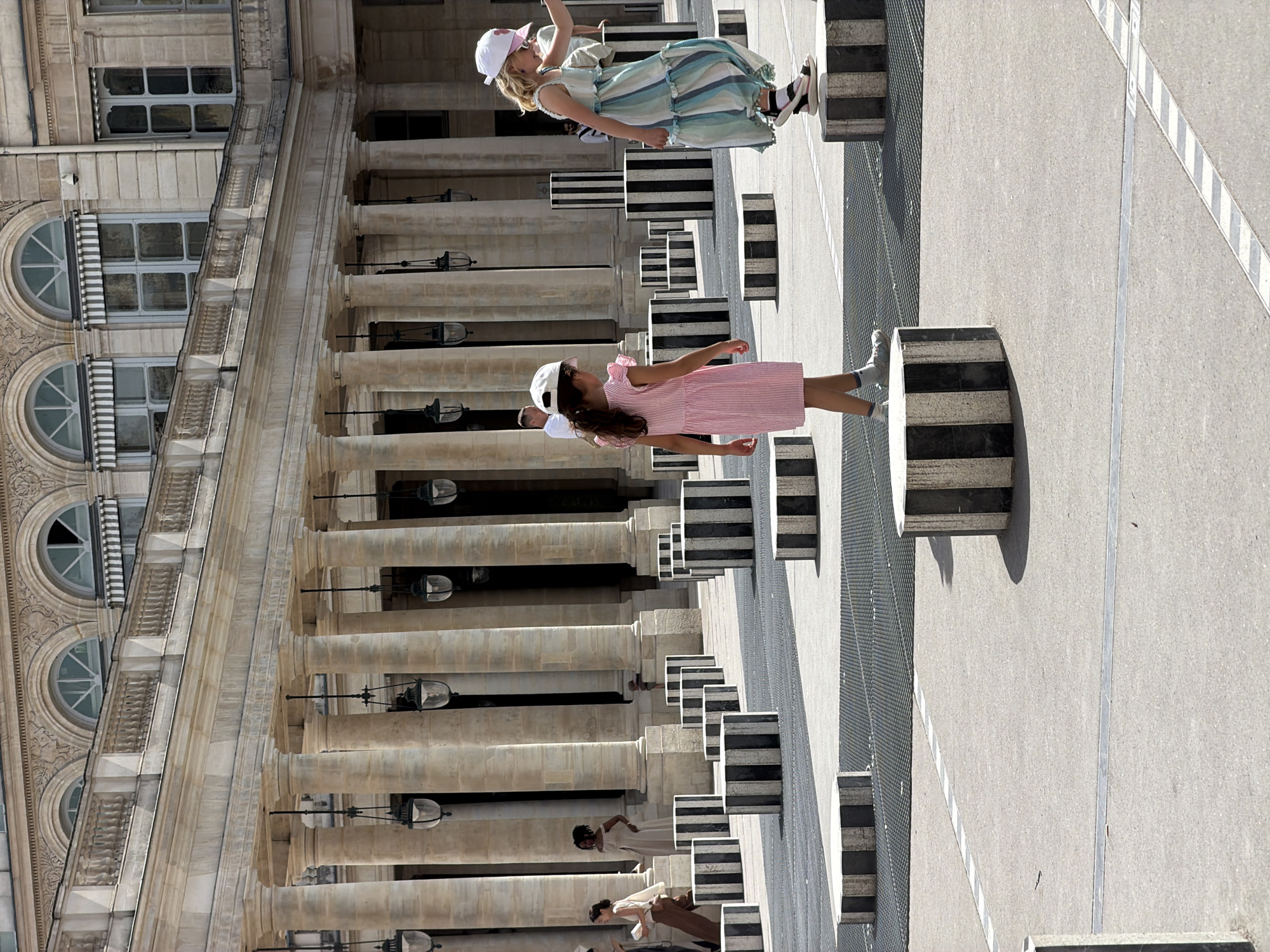 This is a medium shot photograph taken outdoors in a courtyard in Paris, France. The image captures a sunny day with bright lighting and harsh shadows. In the foreground, two young girls are walking on black and white striped cylindrical pedestals. The girl on the left wears a pink striped dress and white cap, while the girl on the right is in a light blue and white striped dress and a white cap. Behind them, a colonnade of large, ornate stone columns with hanging lanterns stretches across the background, supporting a classical architectural facade with arched windows and awnings. Numerous other black and white striped pedestals are scattered throughout the courtyard, some in the mid-ground and others further back. Several adult figures are visible in the background, appearing to be walking or standing near the colonnade, including a woman in a beige outfit and a woman in a light-colored dress. The ground is paved with a light-colored material, with sections of dark metal grating visible. The overall scene suggests a public space, likely a garden or courtyard within a historical Parisian landmark.