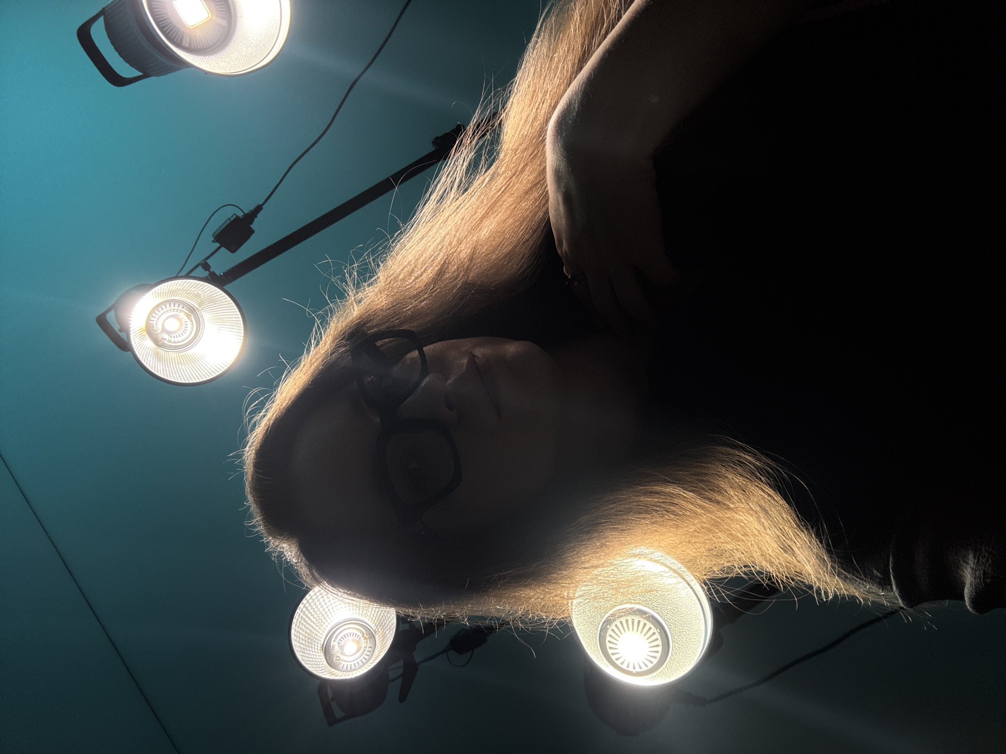 The image shows a person lying on their back looking up at four studio lights. The person has long, light-colored hair and is wearing glasses. The background is a teal color. The setting cannot be determined to be Chaikovsky, Russia, from the visual data alone.