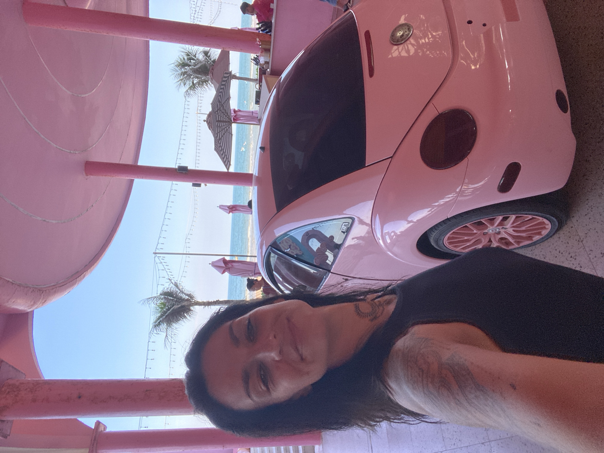 A woman with dark hair and tattoos on her arm and neck smiles at the camera, positioned on the left side of the frame. She is wearing a black tank top. To her right is a pink Volkswagen Beetle, parked under a pink, open-air structure. The structure has pink pillars and a curved pink roof. In the background, beyond the car and structure, is the ocean under a clear blue sky. There are palm trees and beach umbrellas visible near the water's edge. Other people and a bar area are also faintly visible in the background. The overall scene suggests a beachside location, likely a resort or cafe, in Tien Thanh Ward, Vietnam.
