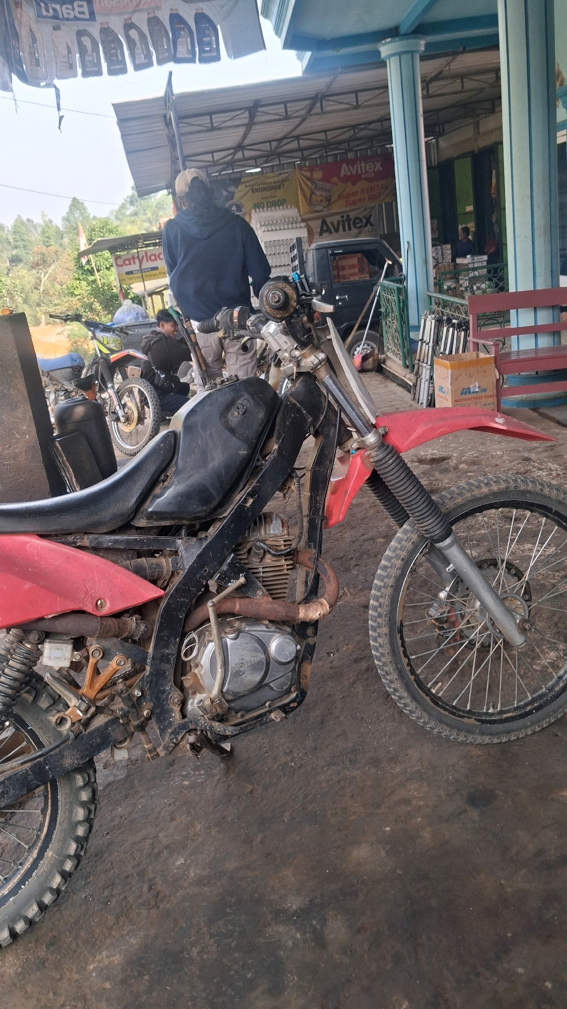 The image depicts a dirt bike with a red and black frame parked on a dirt ground, likely in an open-air workshop or parking area. The motorcycle is positioned in the foreground, facing towards the right of the frame. Its engine and suspension components are visible, showing signs of wear and tear. In the background, there are more motorcycles, a black pickup truck, and shelves stocked with various items, including what appears to be engine oil containers and tires. A person wearing a dark hoodie is seen standing near one of the other motorcycles, and another individual is seated on a motorcycle in the middle ground. The environment suggests a rustic or semi-industrial setting, possibly a motorcycle repair shop or a communal area for vehicle enthusiasts. The overall lighting indicates it's daytime. The location is identified as Karang Sari, Indonesia.