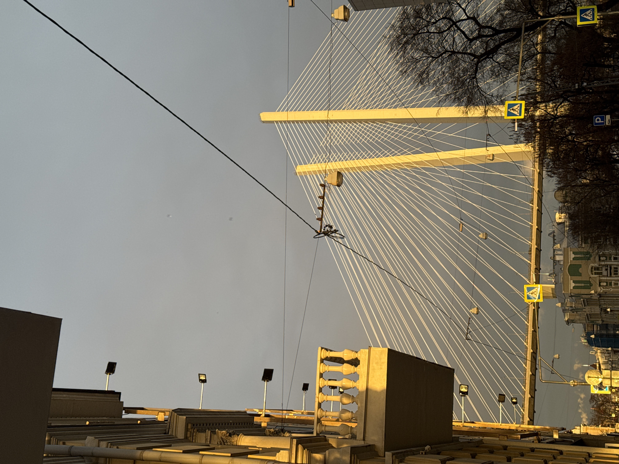 The image depicts the Zolotoy Bridge in Vladivostok, Russia, as seen from below and rotated 90 degrees counter-clockwise. The sky is overcast. Part of a building with multiple spotlights and a decorative facade is visible in the lower part of the image. A power line cuts across the upper left portion of the sky. The bridge's cable-stayed design is prominent, with numerous white cables radiating from two horizontal support beams on the bridge's central tower. Pedestrian crossing signs can be seen on the bridge, positioned vertically due to the image's orientation. Trees and buildings are visible in the background, partly blurred and reflecting the surrounding light.