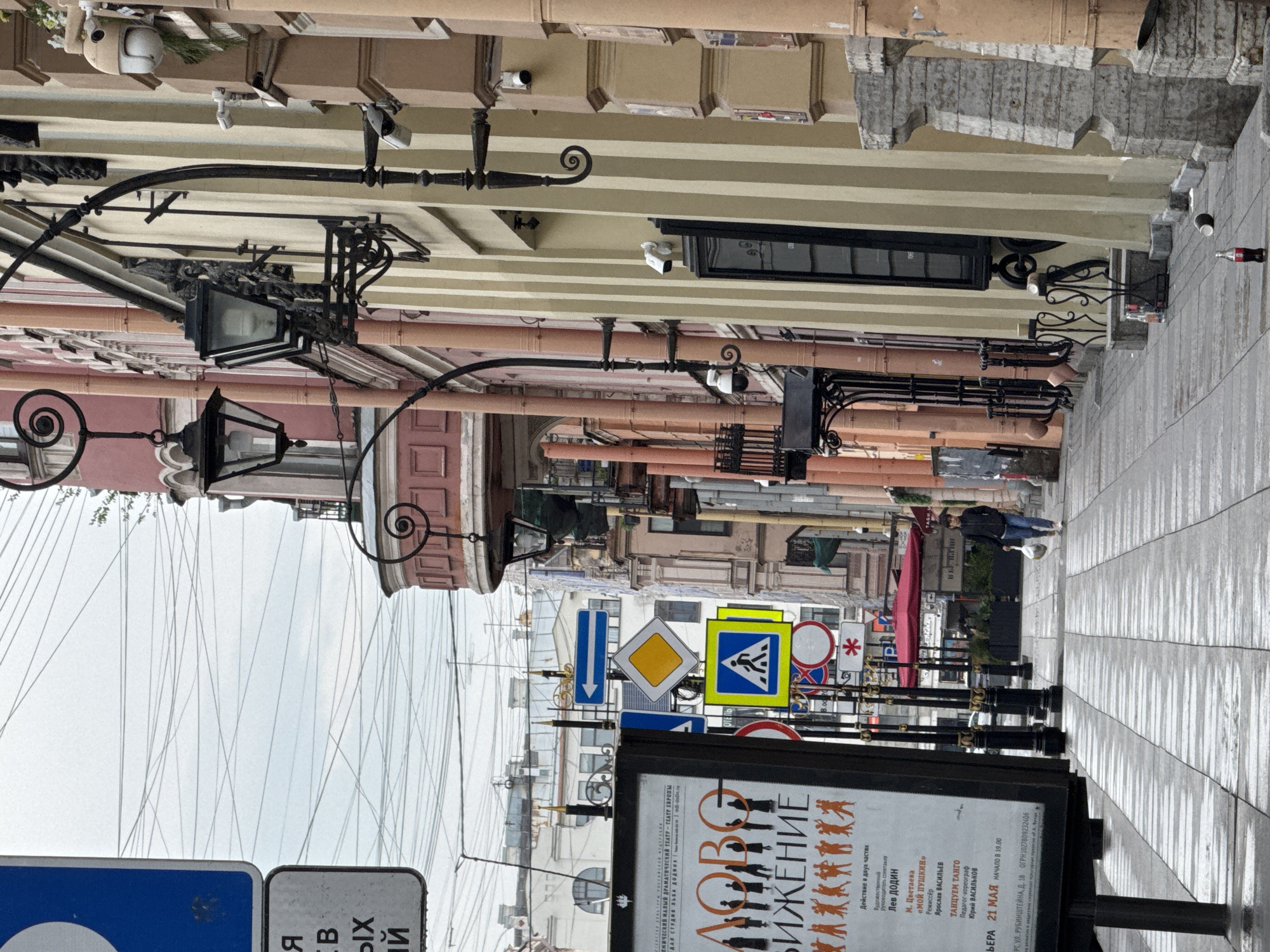 Here is a description of the image:
The image captures a street scene in Saint Petersburg, Russia. The camera is positioned looking down a narrow street lined with buildings on both sides. Overhead, an array of crisscrossing wires are visible. A number of street signs are present, along with a billboard advertising a performance titled "Движение (Movement)" at the Malaya Drama Theatre. The poster features text in Russian with information about the play, playwrights, and performers.
The buildings are characterized by varying architectural styles, with facades displaying different colors and levels of ornamentation. Wall-mounted street lamps with ornate wrought iron details are visible along the buildings. The street pavement is made of stone tiles and appears damp, possibly from recent rain. A person is walking in the distance along the street.