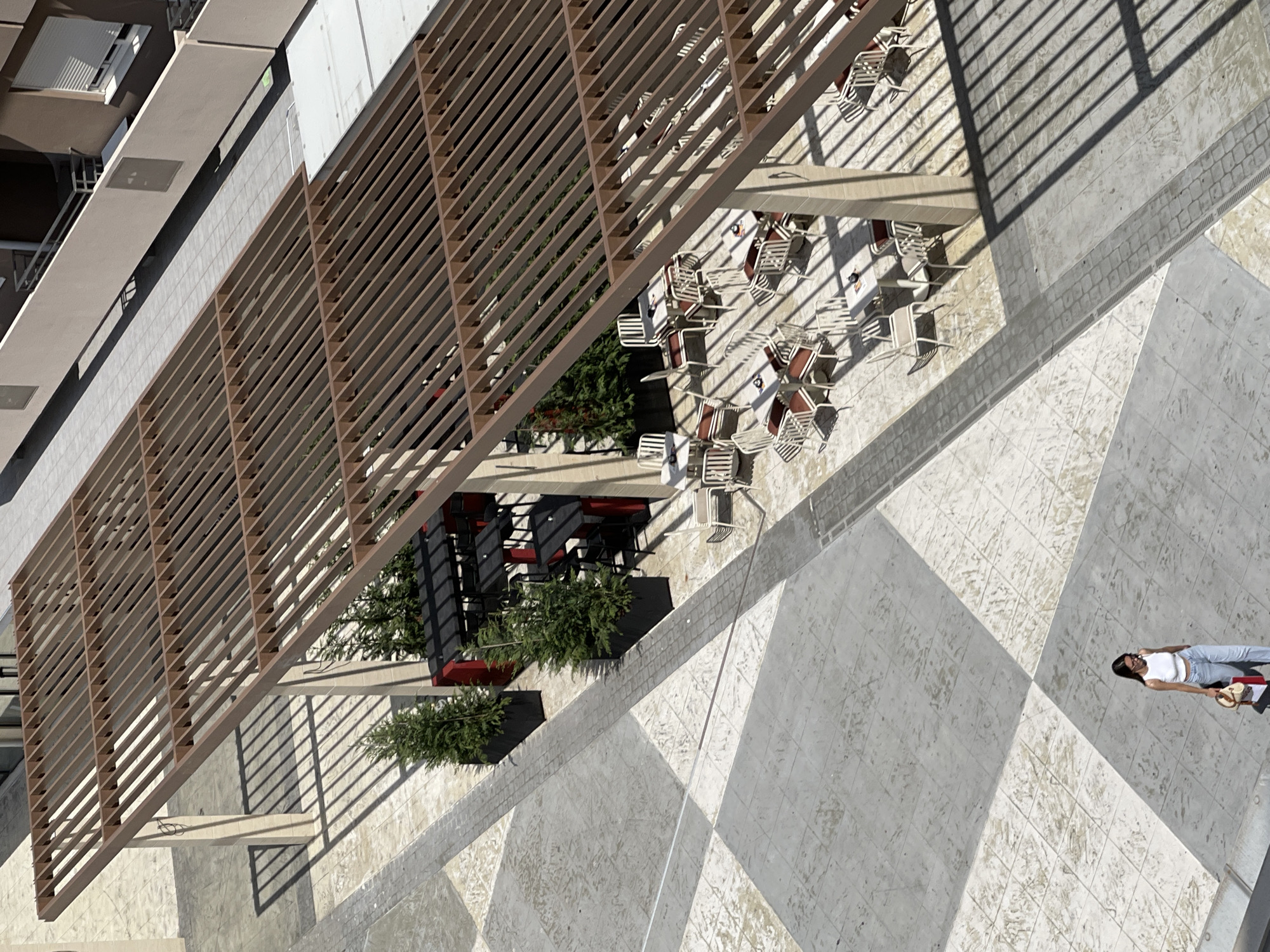 The overhead shot captures an outdoor seating area in Podgorica, Montenegro. A woman wearing a white tank top and blue jeans walks across a paved courtyard with a checkered pattern. She carries a bag. The courtyard is partially covered by a modern wooden pergola, casting striped shadows. Underneath the pergola, several tables and chairs are arranged, suggesting a cafe or restaurant setting. Some tables are empty, while others are set with place settings. Potted evergreen trees are placed strategically near some of the tables. In the background, a building with balconies and windows is visible. The lighting suggests a sunny day.