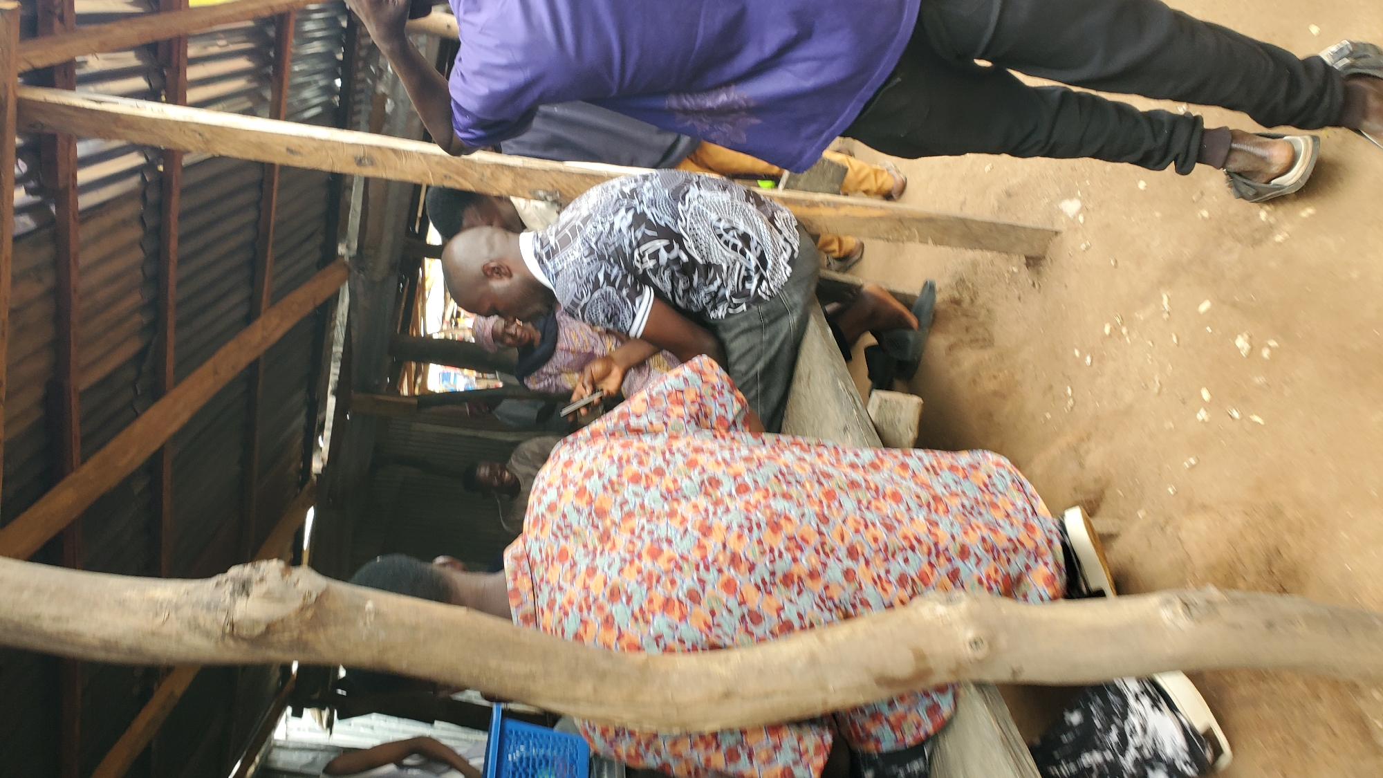 A group of people are gathered under a makeshift structure with a corrugated metal roof supported by wooden beams and poles. In the foreground, a person wearing a vibrant, patterned shirt faces away from the camera. To their right, another individual wearing a black and white patterned shirt is seated, looking at a smartphone. Behind them, a woman in a headscarf and patterned clothing appears to be looking at the person with the phone. Further back, other individuals are visible in the dim light of the structure, some appearing to be talking and laughing. The ground is dirt, with scattered debris. The structure appears to be an outdoor market stall or gathering place. The scene is located in Jalingo, Nigeria.