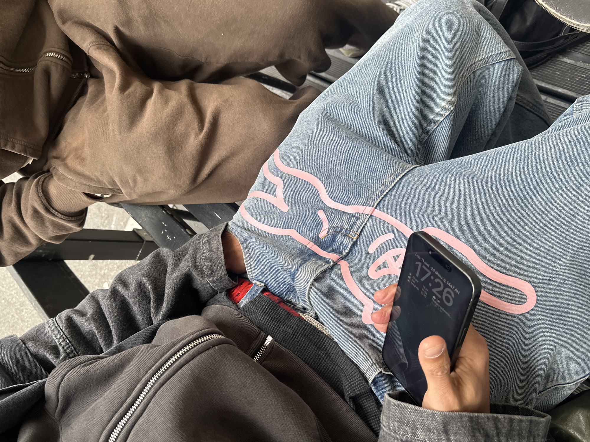 A close-up shot shows a person seated, holding a smartphone in their right hand. The phone screen displays the date and time as "Lun. 11 mai 24 dhou. q. 1447 AH 17:26." The person is wearing blue jeans with a pink graphic design on the upper thigh and a gray zipper hoodie. Part of a brown hoodie is also visible to the left, suggesting another person is sitting nearby. A black bag strap crosses the person's lap. The location is not identifiable as being in Paris, France.