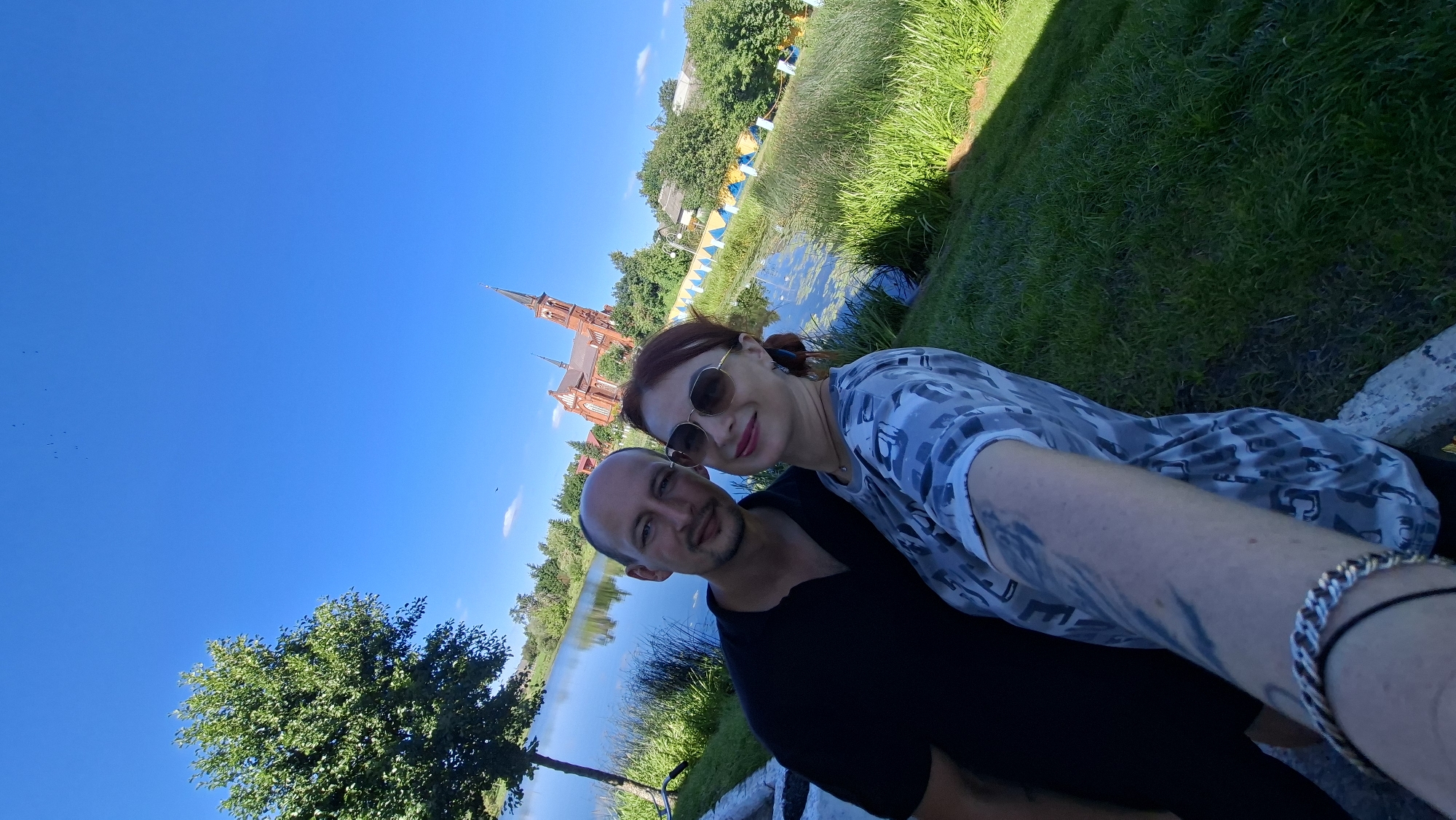 A man and a woman are taking a selfie by a body of water. The man is wearing a black polo shirt, and the woman is wearing a grey and white camouflage t-shirt and sunglasses. In the background, there is a red brick church with a tall steeple, a tree, and lush green grass. The sky is clear and blue. The location is Pastavy, Belarus.